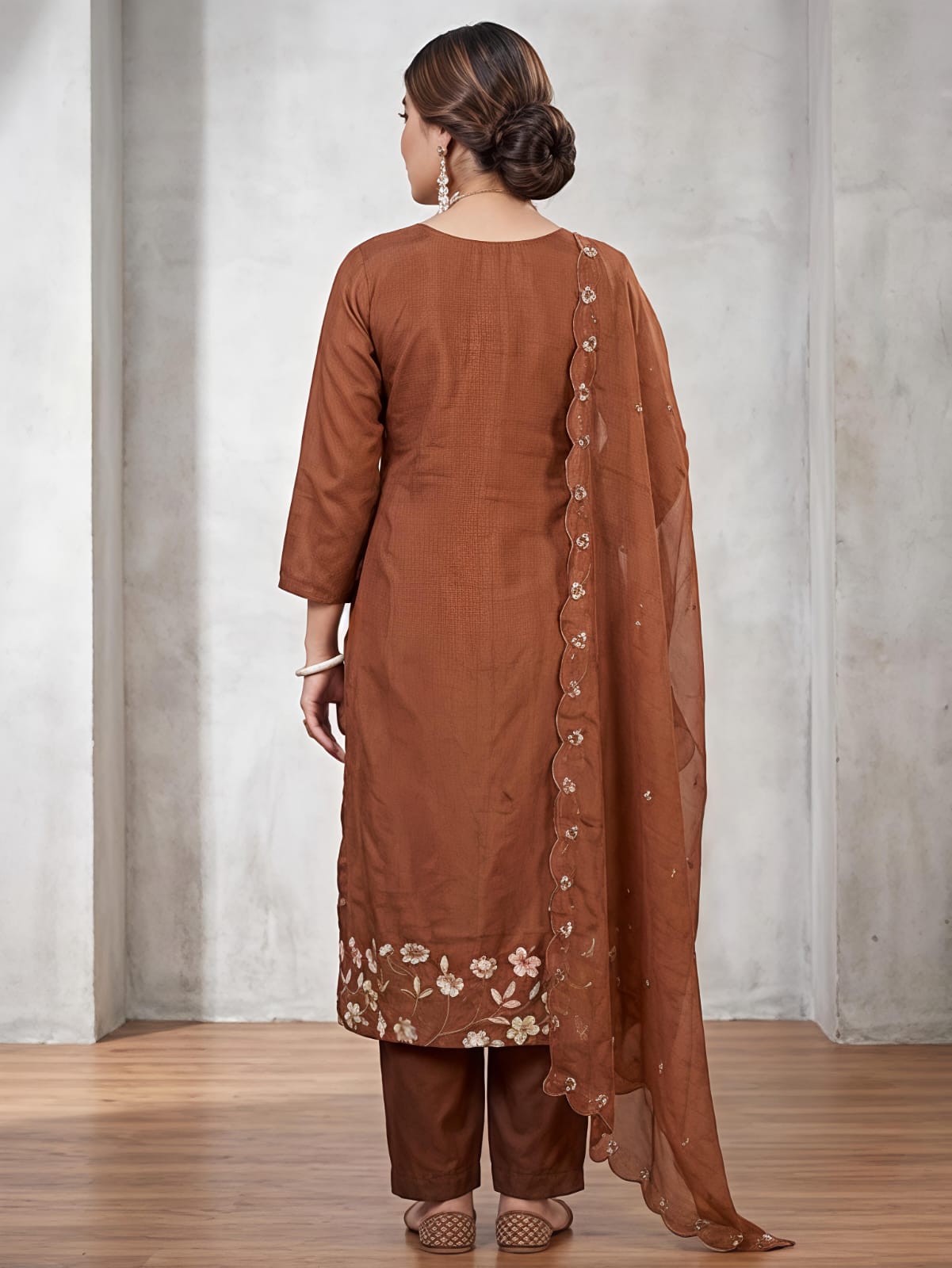 A woman models a brown traditional Indian salwar kameez suit set, featuring a brown kurta with floral embroidery at the hem, matching brown trousers, and a sheer brown dupatta with decorative buttons down the side, set against a gray concrete wall background.