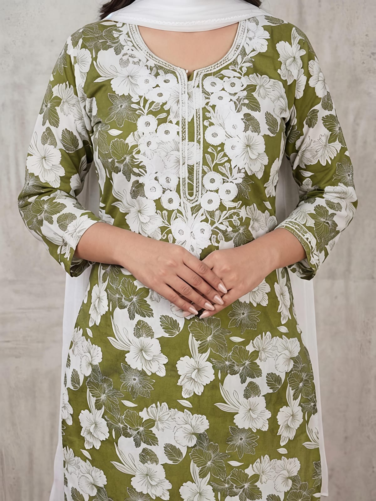 A woman modeling a green and white floral printed kurta set with matching pants and a white dupatta, standing in front of a textured gray backdrop.