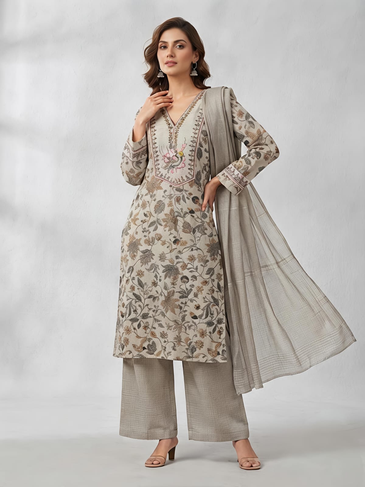 A woman models a traditional three-piece salwar kameez suit in a light beige and gray floral print, featuring a long-sleeved embroidered kameez top, wide-leg trousers, and a matching sheer dupatta, set against a neutral gray studio backdrop.