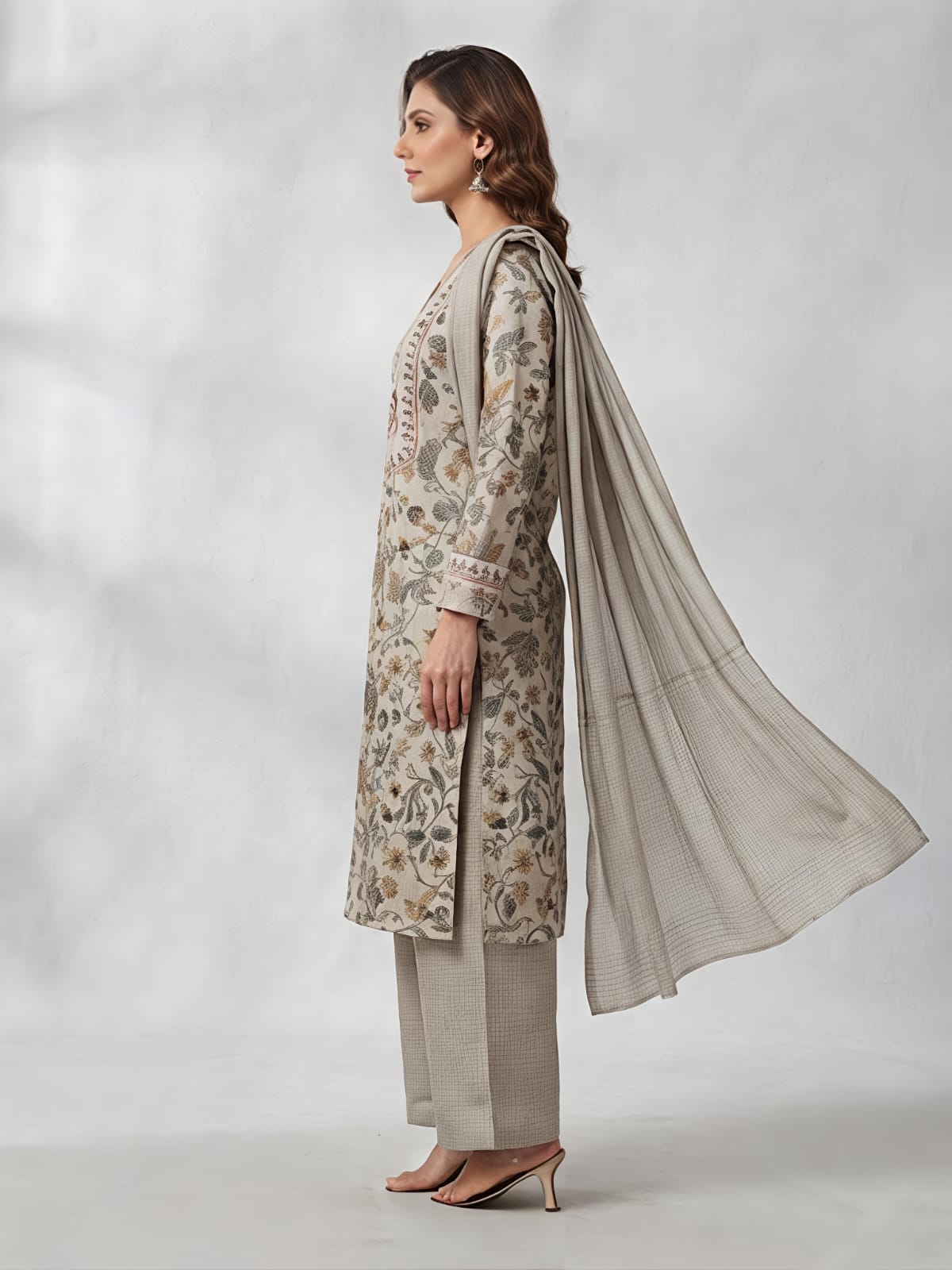 A woman models a traditional three-piece salwar kameez suit in a light beige and gray floral print, featuring a long-sleeved embroidered kameez top, wide-leg trousers, and a matching sheer dupatta, set against a neutral gray studio backdrop.
