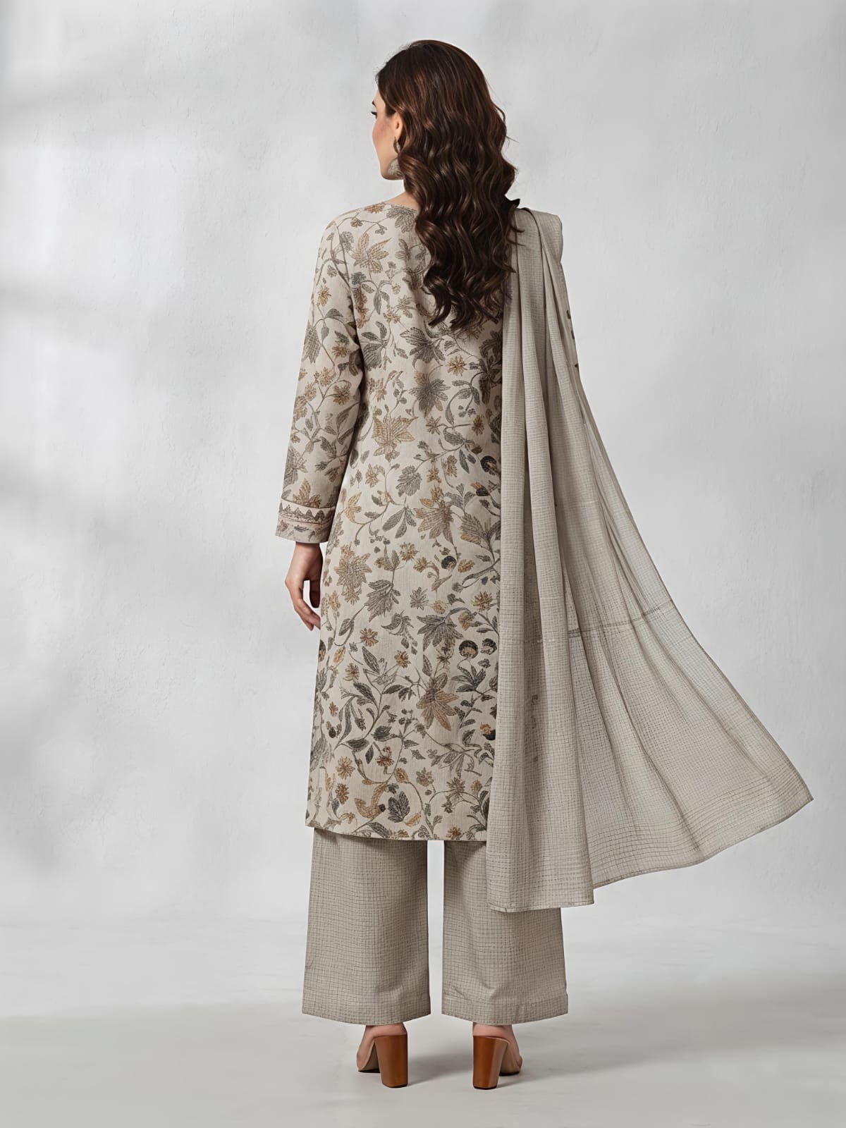 A woman models a traditional three-piece salwar kameez suit in a light beige and gray floral print, featuring a long-sleeved embroidered kameez top, wide-leg trousers, and a matching sheer dupatta, set against a neutral gray studio backdrop.