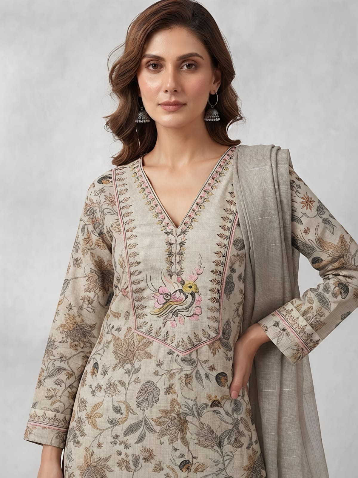 A woman models a traditional three-piece salwar kameez suit in a light beige and gray floral print, featuring a long-sleeved embroidered kameez top, wide-leg trousers, and a matching sheer dupatta, set against a neutral gray studio backdrop.
