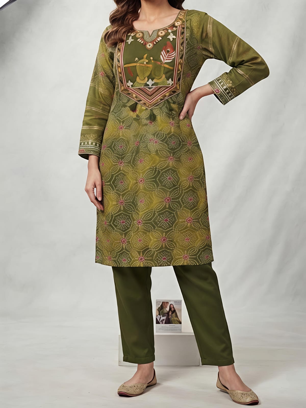 A woman models an olive green salwar kameez suit with a matching dupatta. The kurta features a yellow-gold base with intricate green and pink floral patterns, and a detailed embroidered neckline.