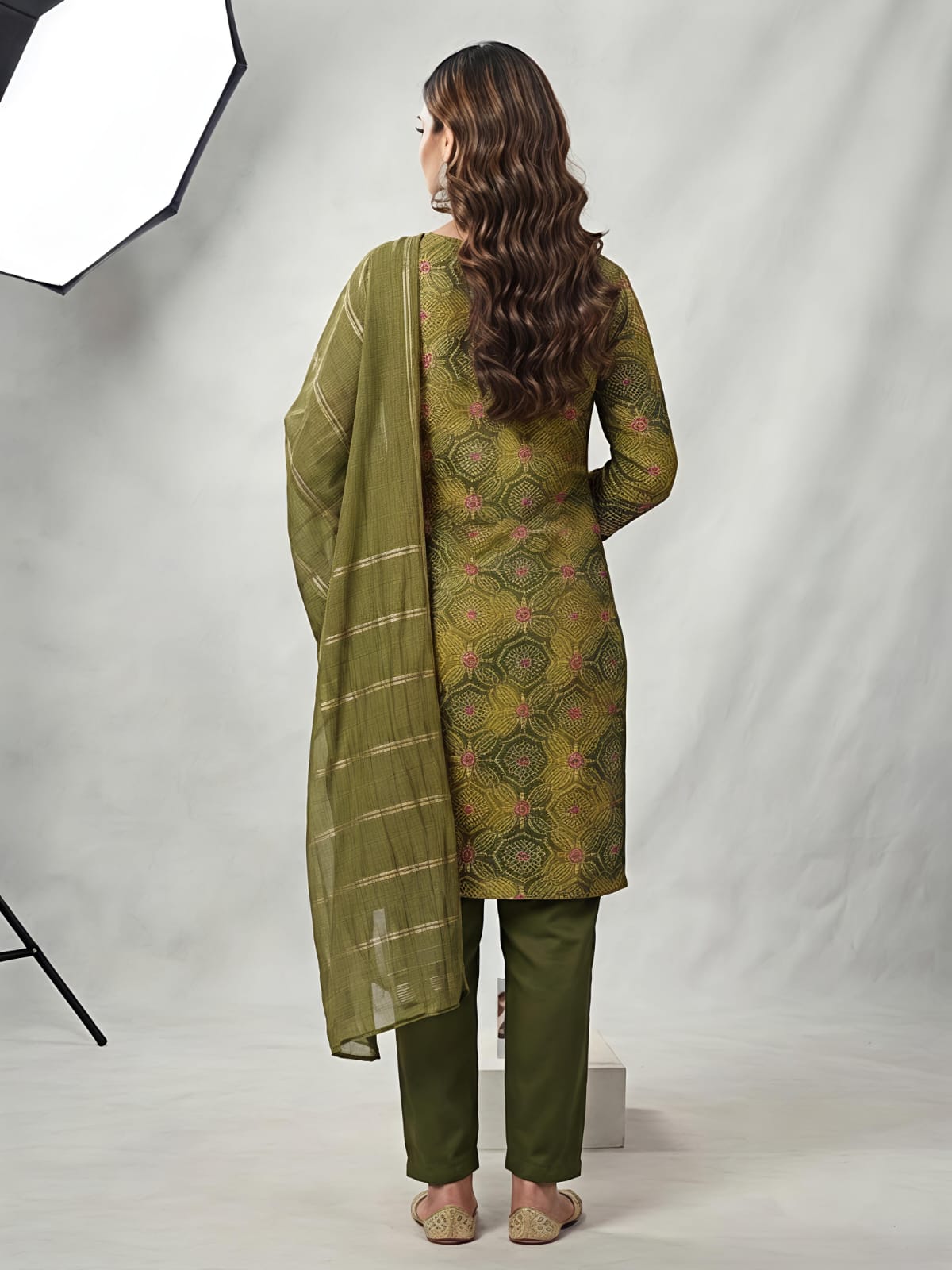 A woman models an olive green salwar kameez suit with a matching dupatta. The kurta features a yellow-gold base with intricate green and pink floral patterns, and a detailed embroidered neckline.