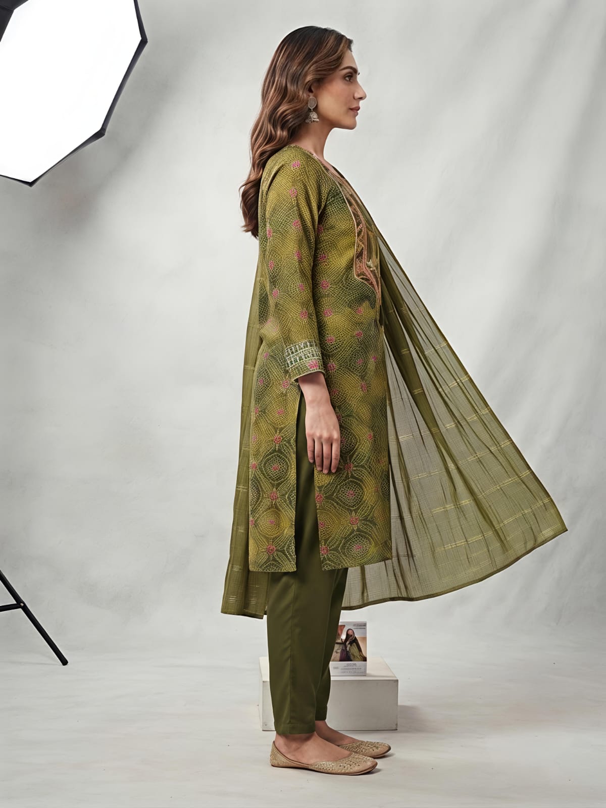 A woman models an olive green salwar kameez suit with a matching dupatta. The kurta features a yellow-gold base with intricate green and pink floral patterns, and a detailed embroidered neckline.