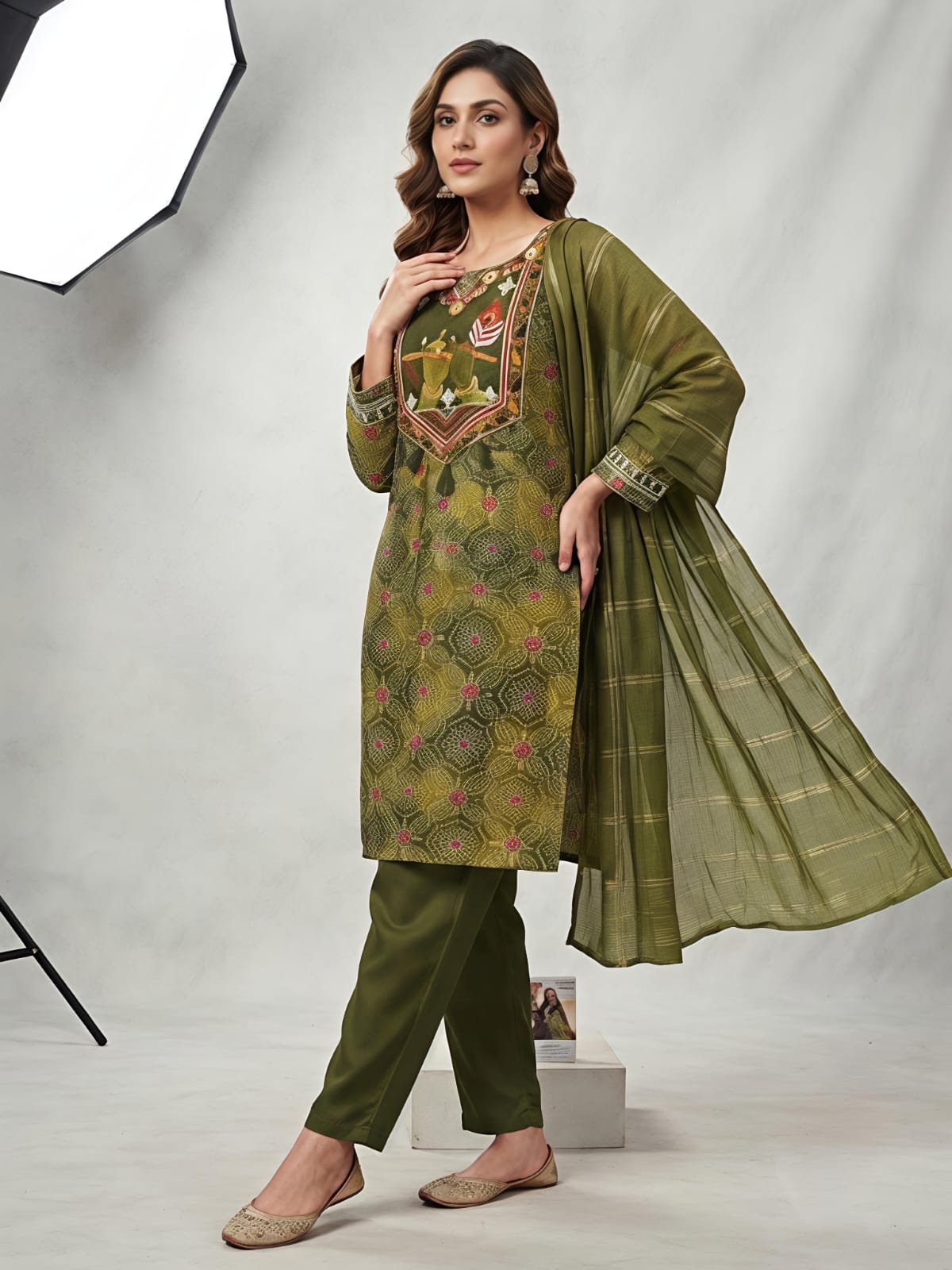 A woman models an olive green salwar kameez suit with a matching dupatta. The kurta features a yellow-gold base with intricate green and pink floral patterns, and a detailed embroidered neckline.