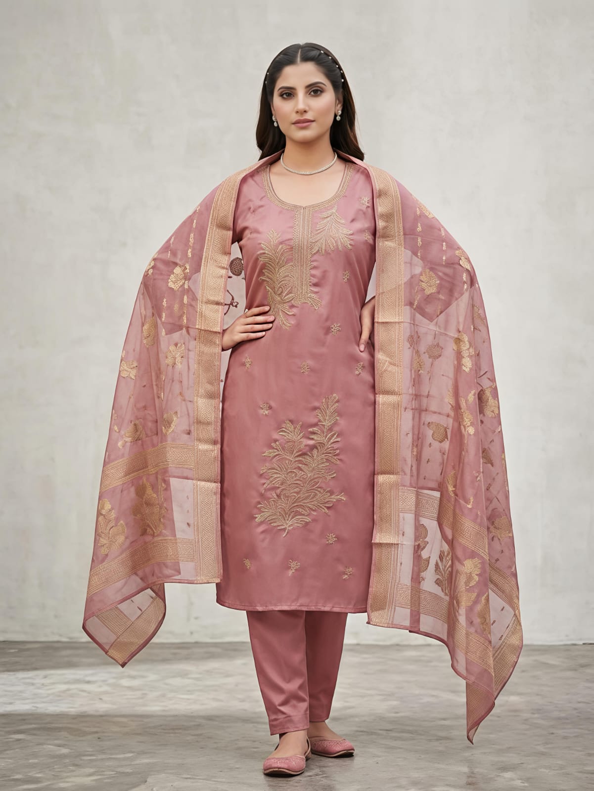 A woman modeling a dusty pink traditional salwar kameez suit with gold floral embroidery, matching trousers, and a sheer dupatta draped over her shoulders, set against a neutral gray studio background.
