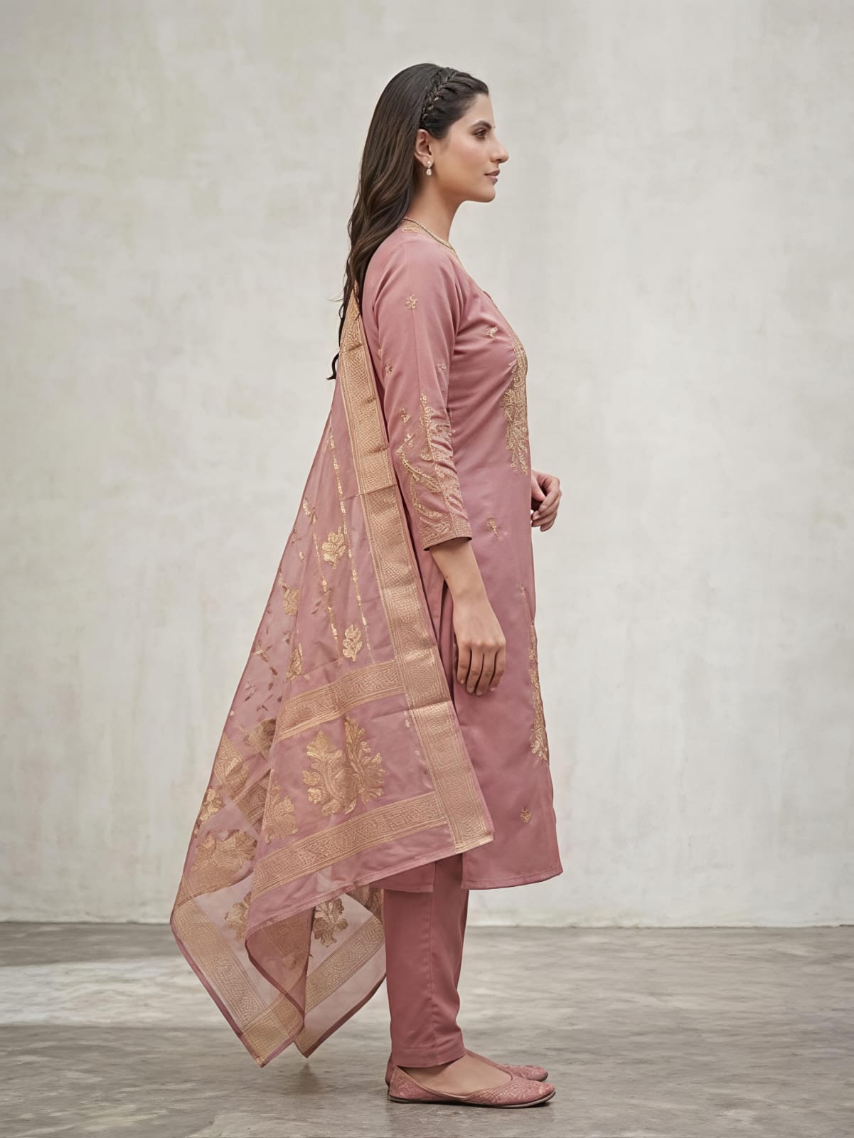 A woman modeling a dusty pink traditional salwar kameez suit with gold floral embroidery, matching trousers, and a sheer dupatta draped over her shoulders, set against a neutral gray studio background.