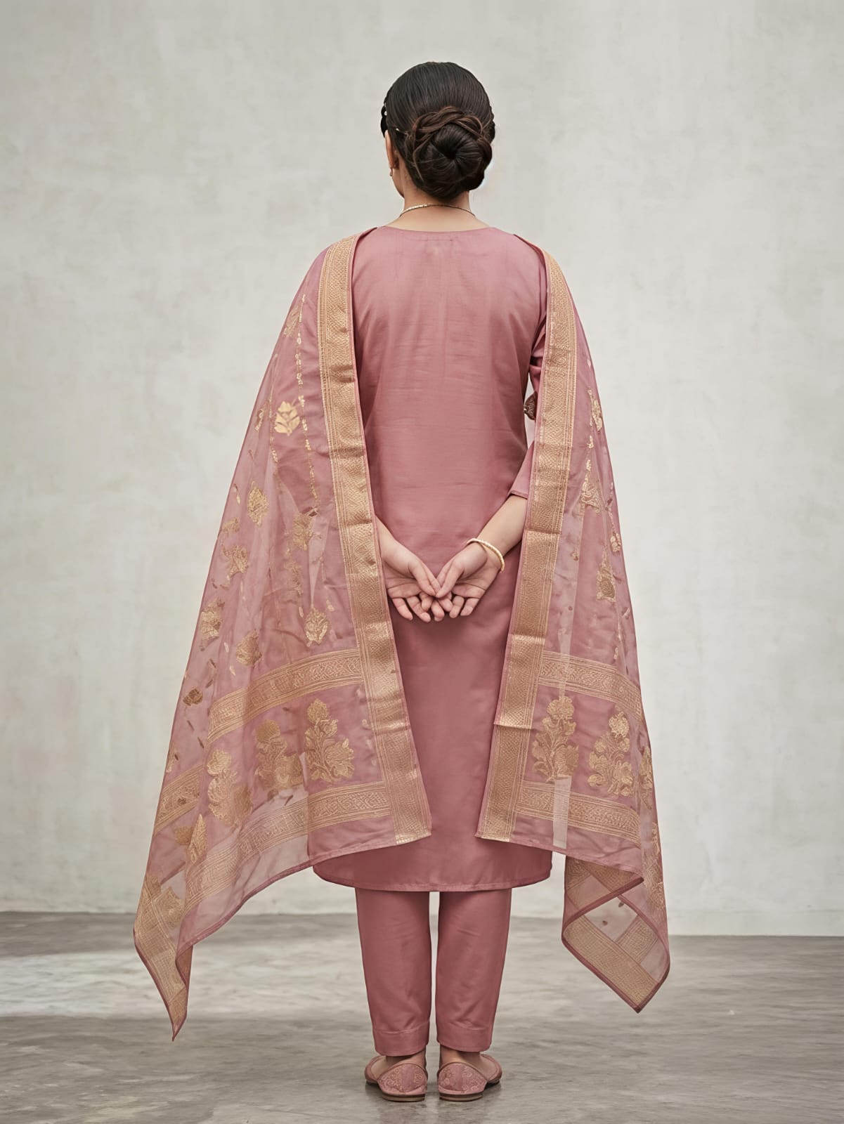 A woman modeling a dusty pink traditional salwar kameez suit with gold floral embroidery, matching trousers, and a sheer dupatta draped over her shoulders, set against a neutral gray studio background.