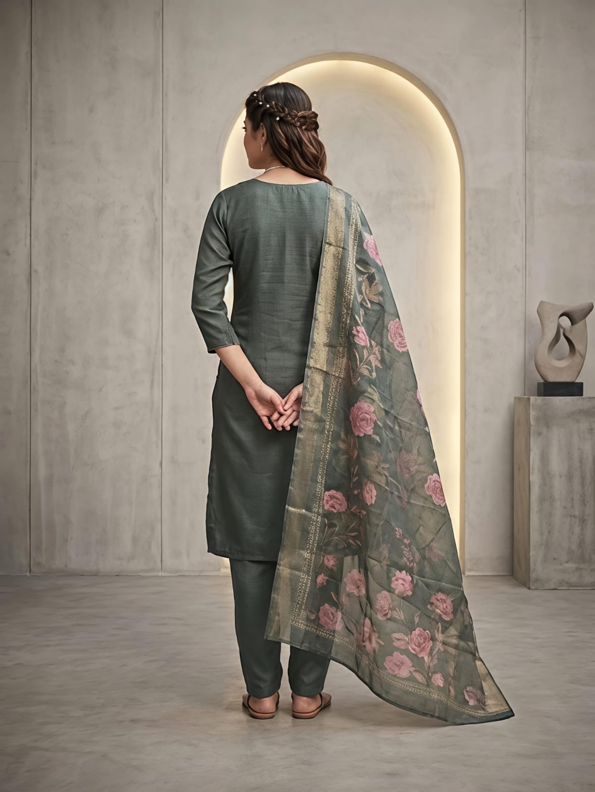 A woman models a three-piece salwar kameez suit in a muted sage green color, featuring floral embroidery on the kurta and a matching printed dupatta with pink flowers, set against a modern concrete archway background.
