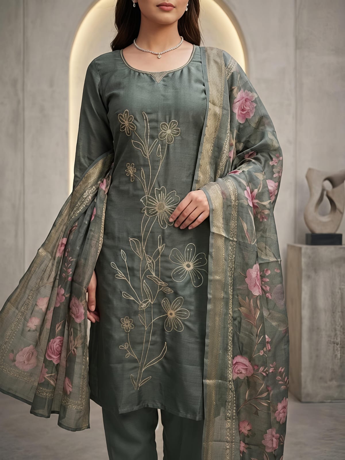 A woman models a three-piece salwar kameez suit in a muted sage green color, featuring floral embroidery on the kurta and a matching printed dupatta with pink flowers, set against a modern concrete archway background.