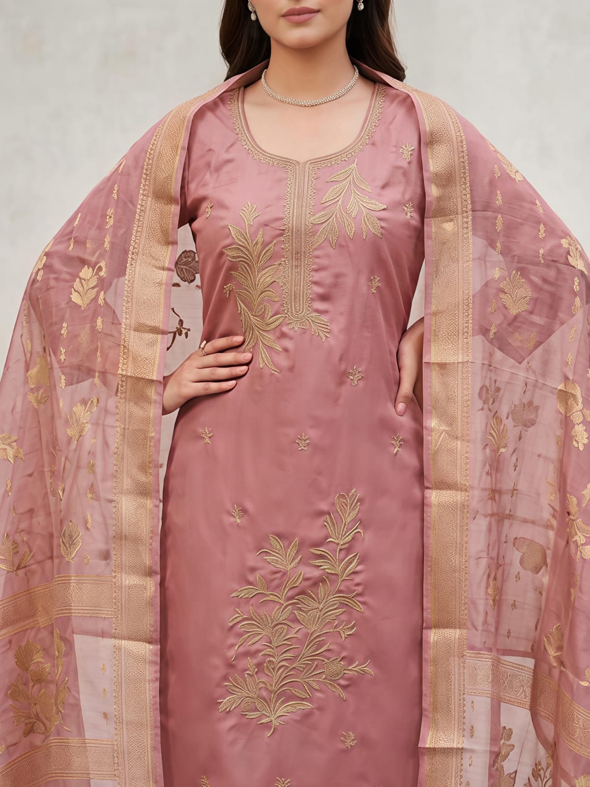 A woman modeling a dusty pink traditional salwar kameez suit with gold floral embroidery, matching trousers, and a sheer dupatta draped over her shoulders, set against a neutral gray studio background.