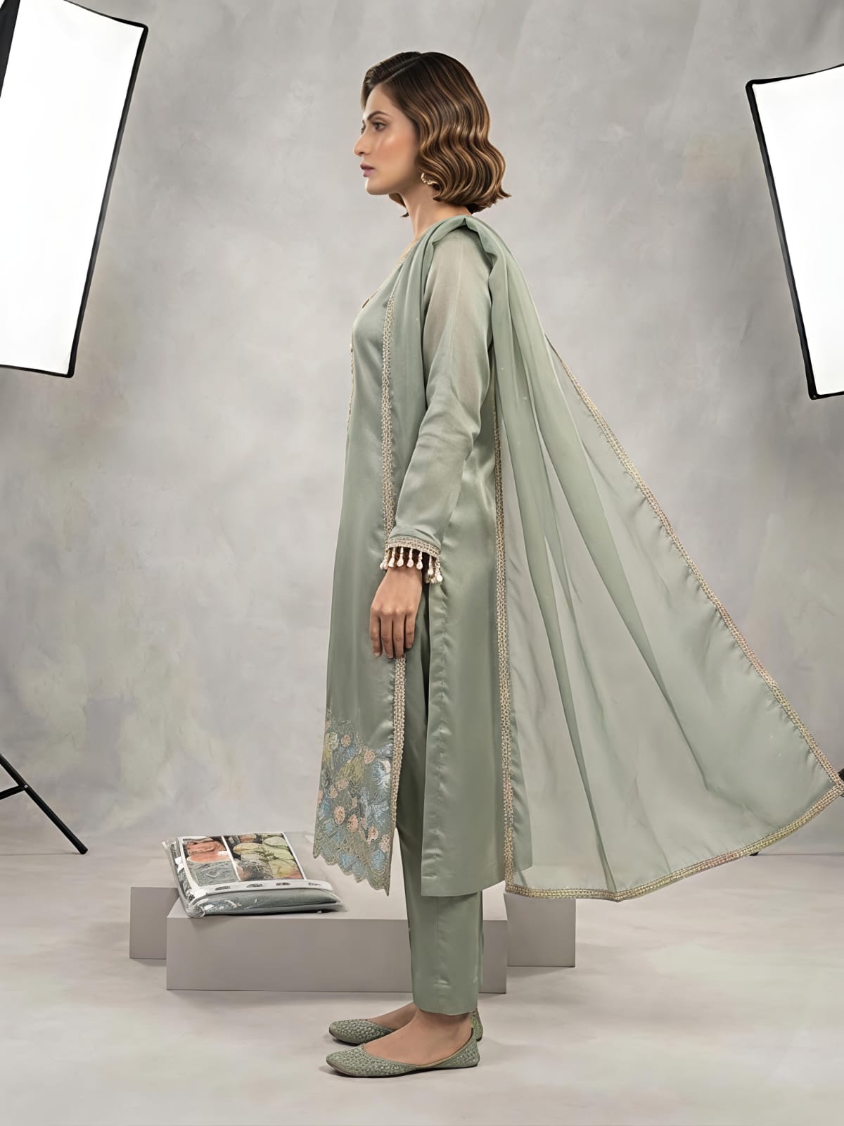 A woman models a light sage green traditional outfit consisting of a long kurta with floral embroidery, matching trousers, and a sheer dupatta, set against a gray studio backdrop with studio lights visible.