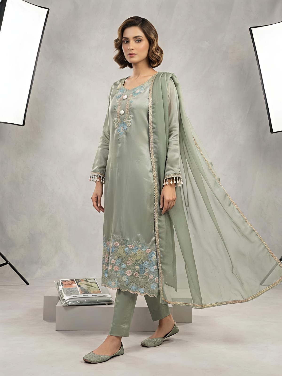 A woman models a light sage green traditional outfit consisting of a long kurta with floral embroidery, matching trousers, and a sheer dupatta, set against a gray studio backdrop with studio lights visible.