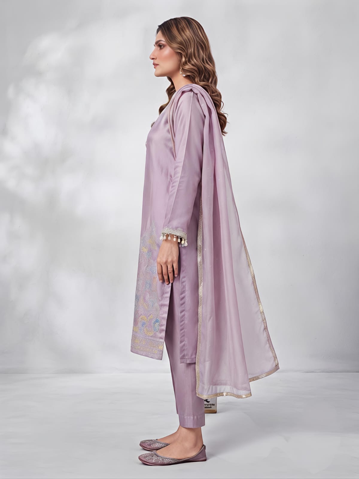 A woman models a three-piece traditional South Asian salwar kameez set in a dusty lavender purple. The set includes a long-sleeved embroidered kameez kurta, matching trousers, and a sheer dupatta.
