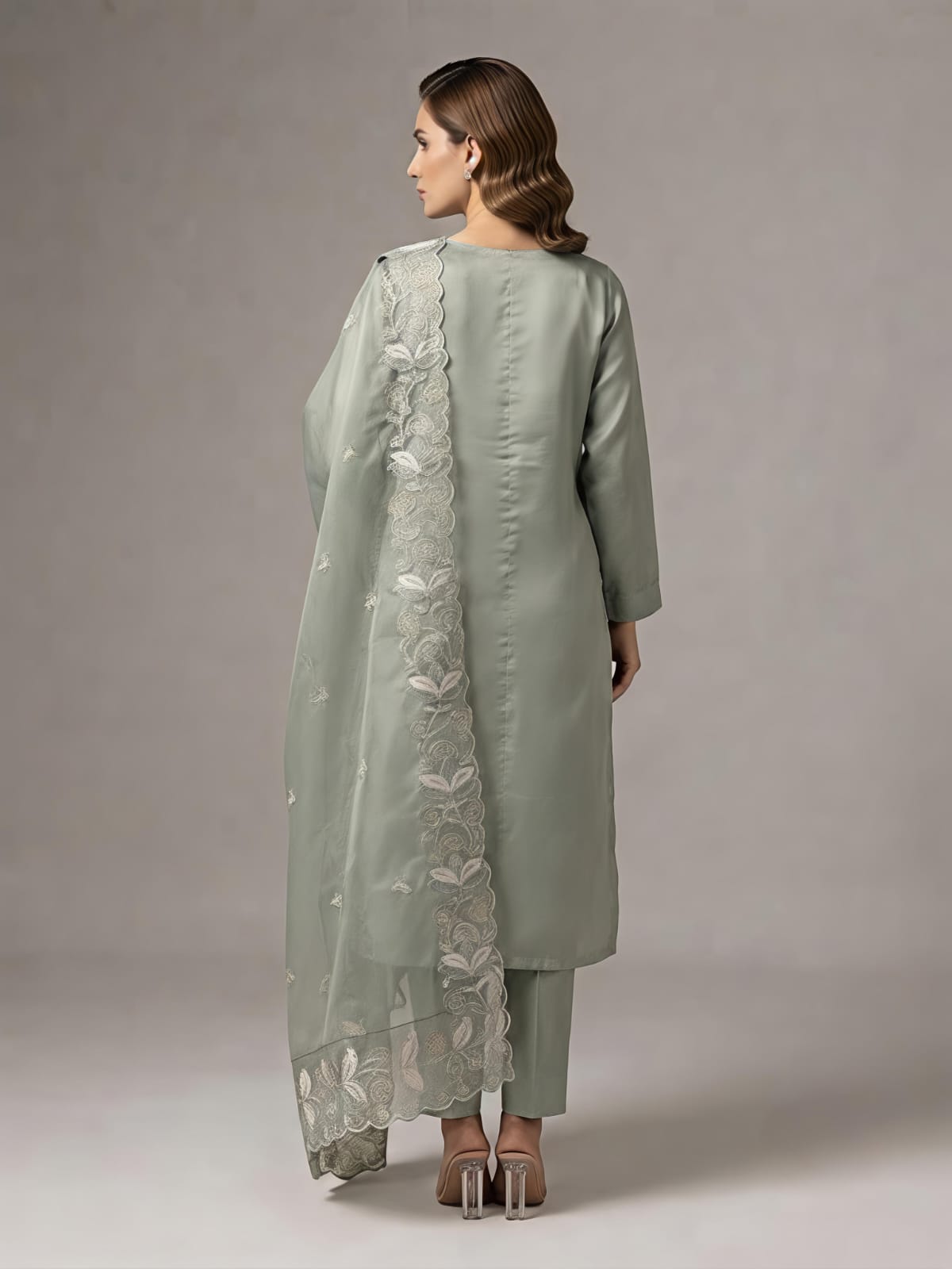 A woman models a sage green traditional Indian outfit consisting of a long kurta with floral embroidery, matching pants, and a sheer dupatta, set against a plain gray studio background.