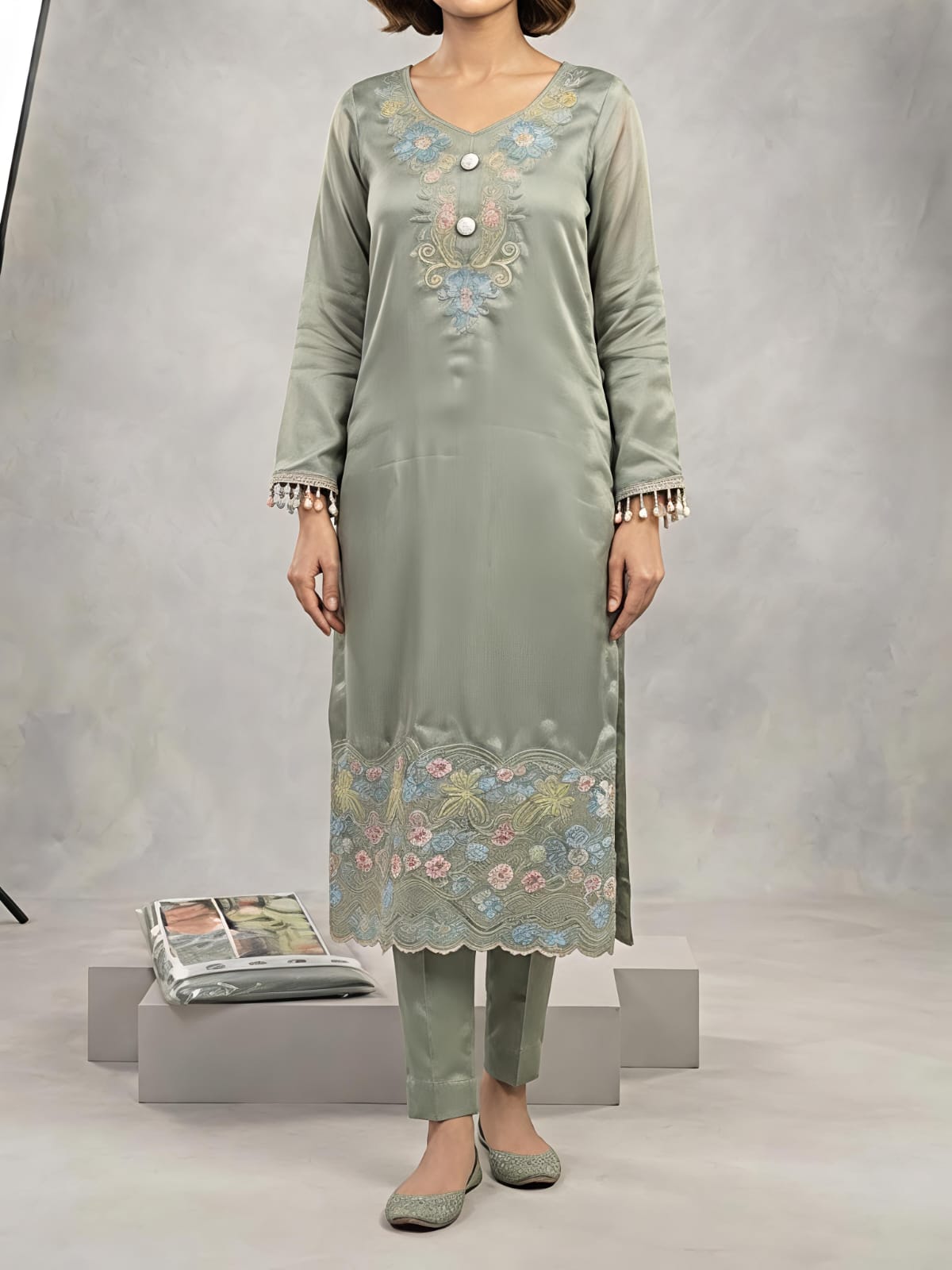 A woman models a light sage green traditional outfit consisting of a long kurta with floral embroidery, matching trousers, and a sheer dupatta, set against a gray studio backdrop with studio lights visible.