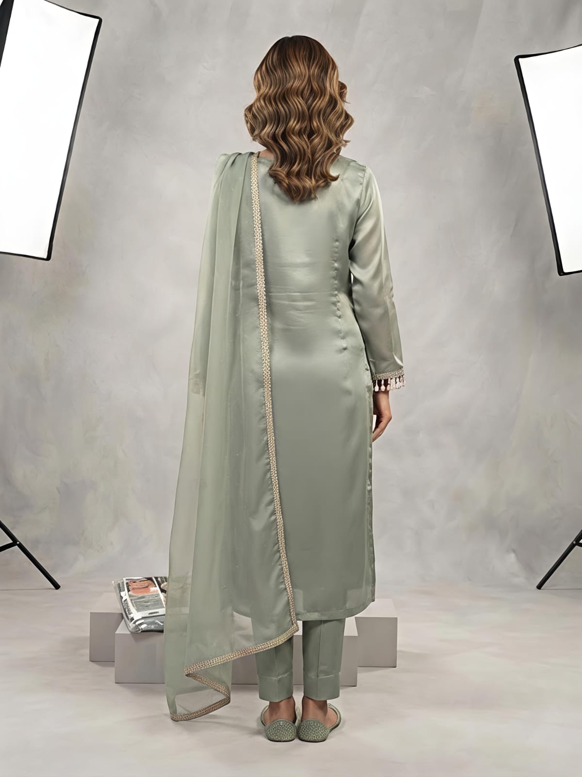 A woman models a light sage green traditional outfit consisting of a long kurta with floral embroidery, matching trousers, and a sheer dupatta, set against a gray studio backdrop with studio lights visible.