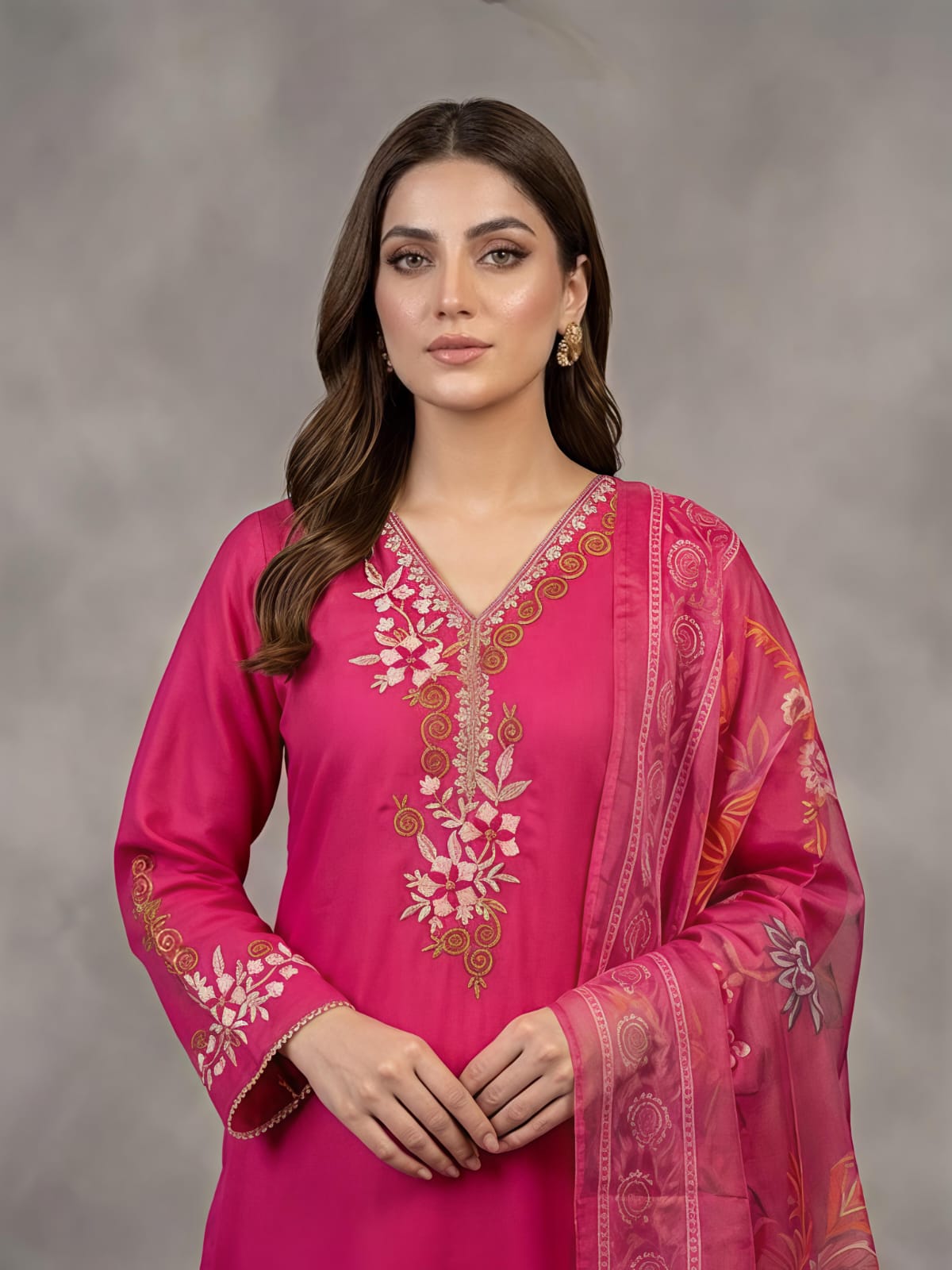 A woman models a bright pink traditional outfit with matching pants and a sheer printed dupatta, standing against a textured gray studio backdrop.