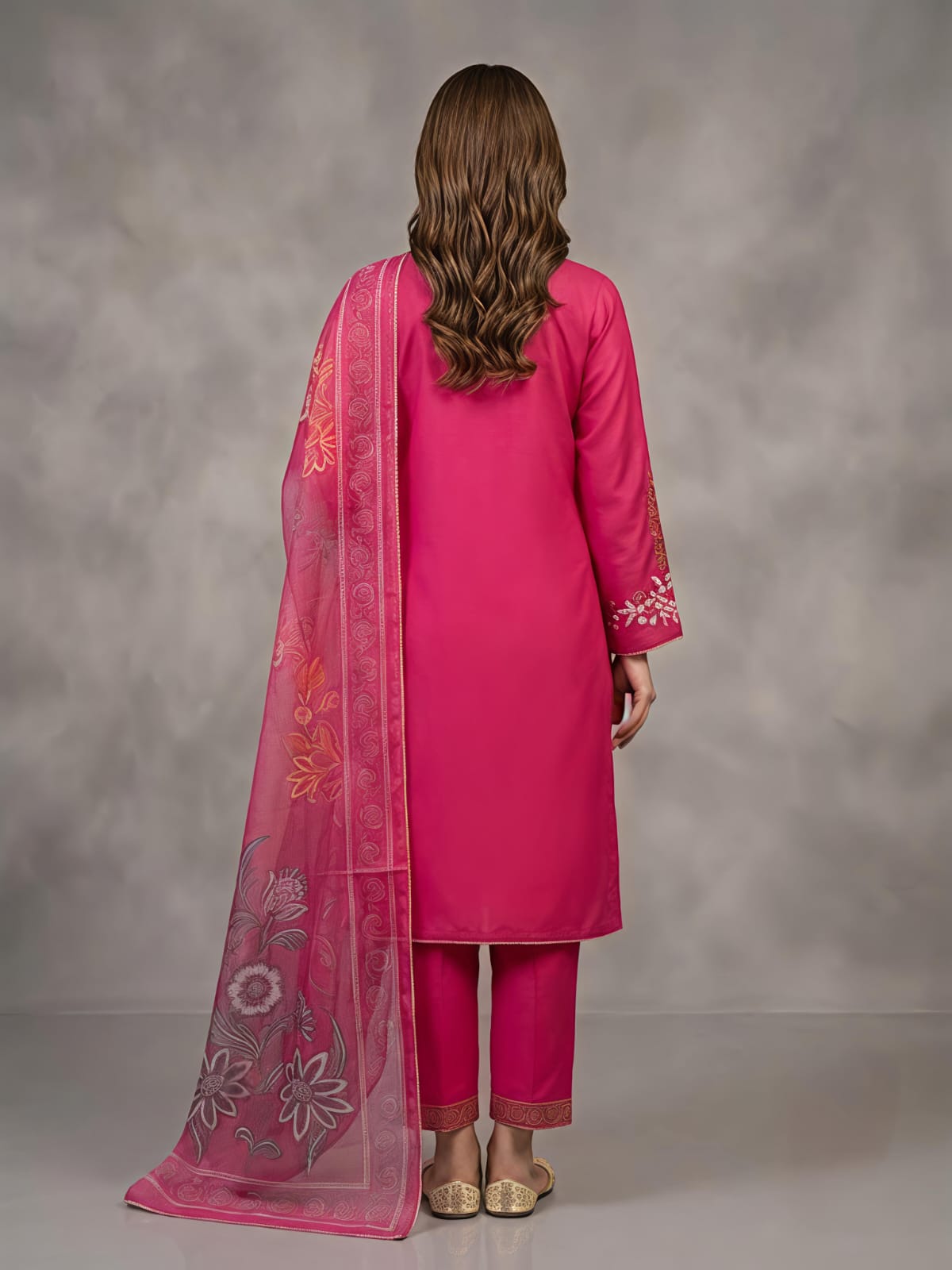 A woman models a bright pink traditional outfit with matching pants and a sheer printed dupatta, standing against a textured gray studio backdrop.