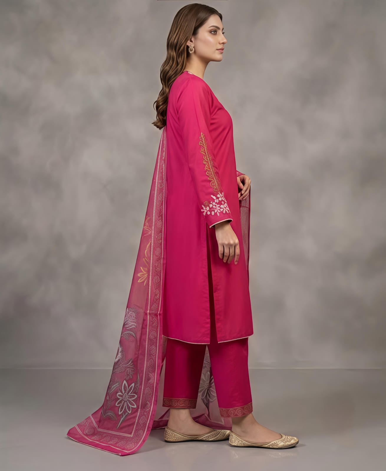 A woman models a bright pink traditional outfit with matching pants and a sheer printed dupatta, standing against a textured gray studio backdrop.