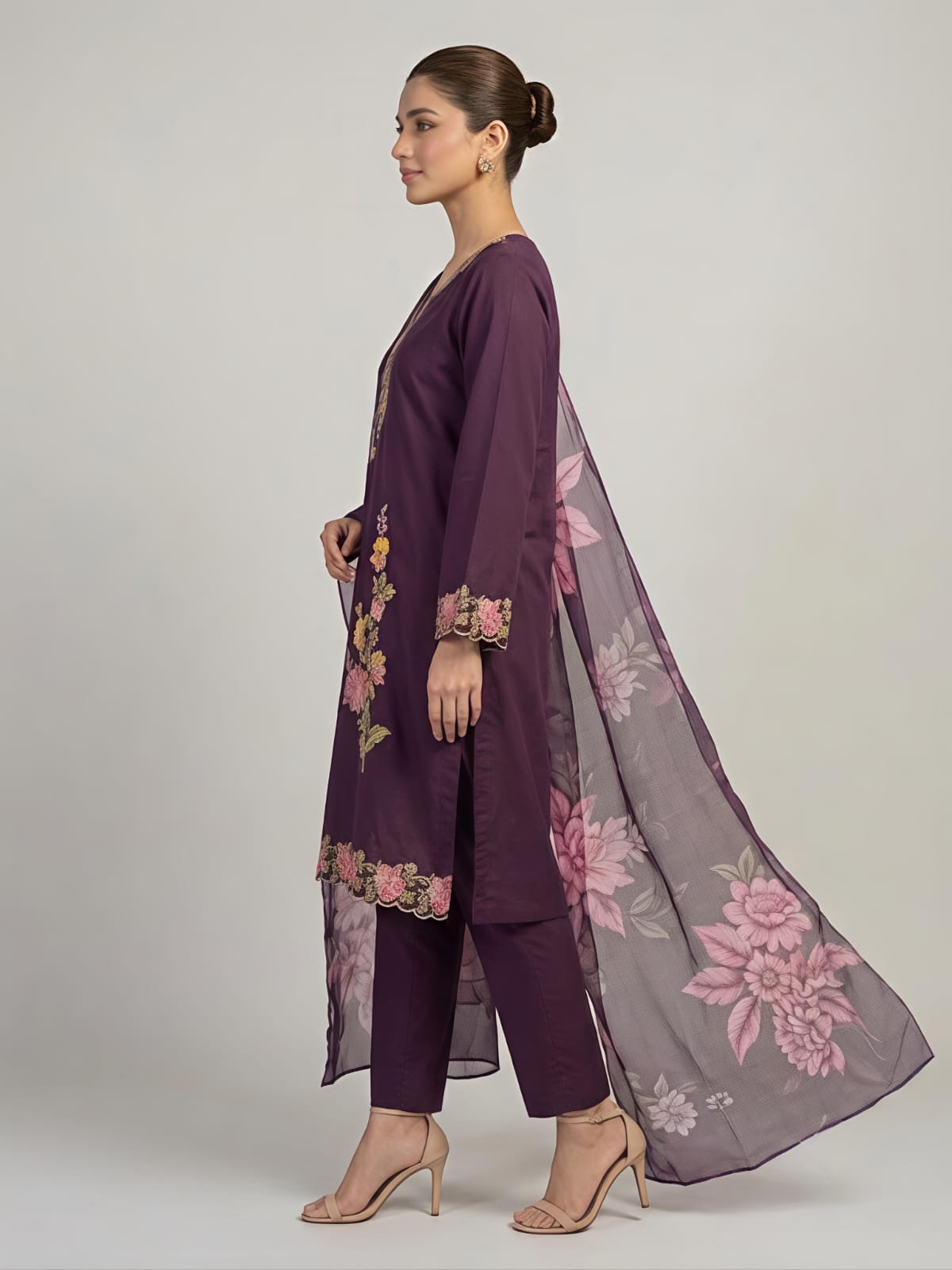 A woman modeling a deep purple three-piece traditional outfit with floral embroidery and a matching sheer dupatta, set against a plain light gray studio background.