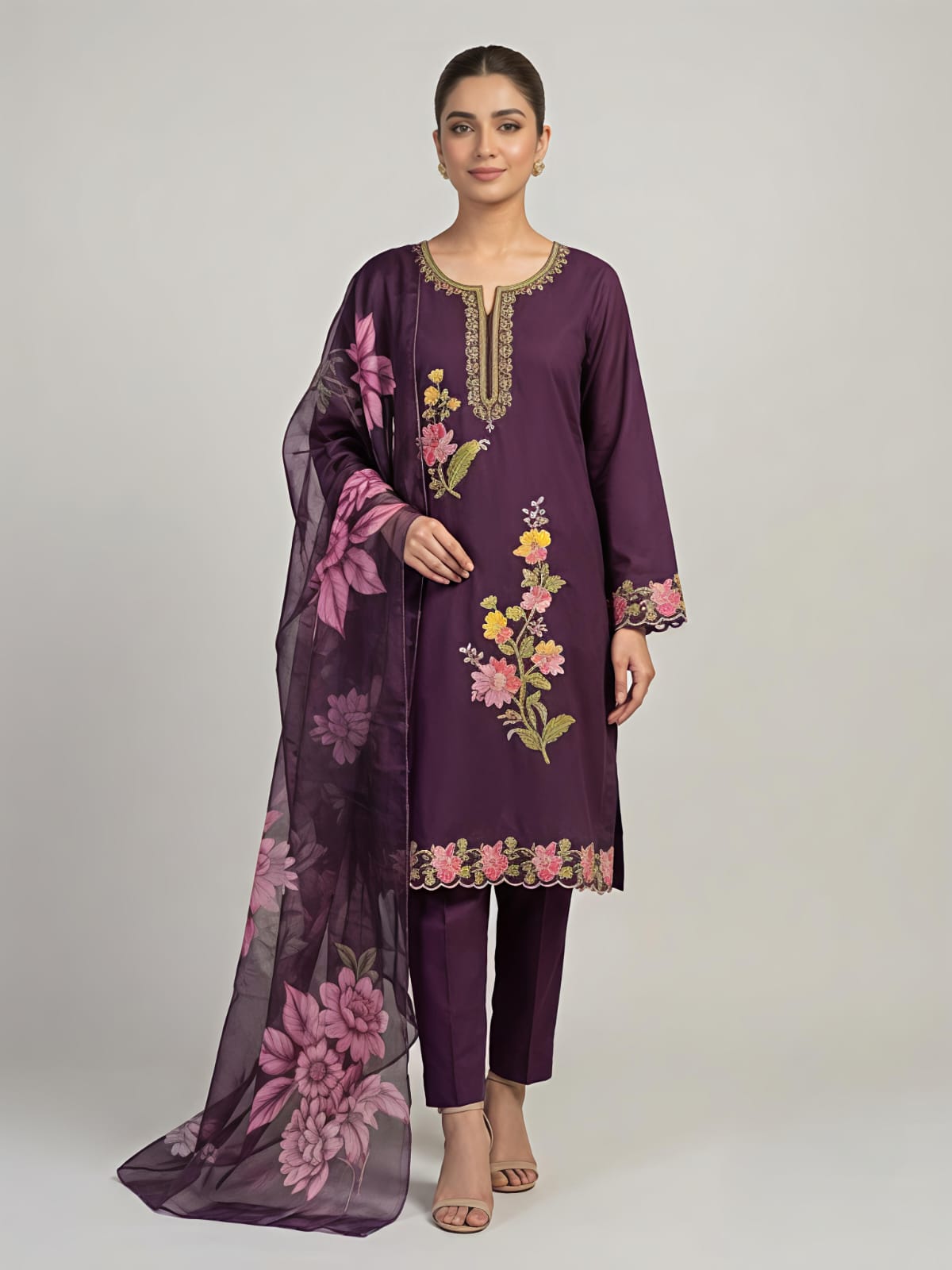 A woman modeling a deep purple three-piece traditional outfit with floral embroidery and a matching sheer dupatta, set against a plain light gray studio background.