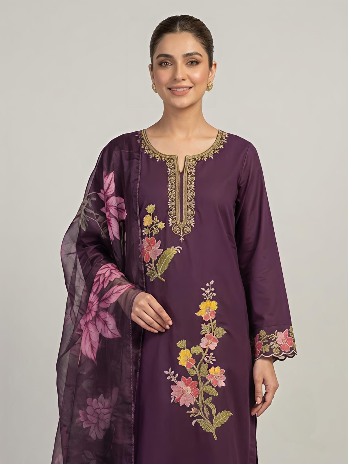 A woman modeling a deep purple three-piece traditional outfit with floral embroidery and a matching sheer dupatta, set against a plain light gray studio background.