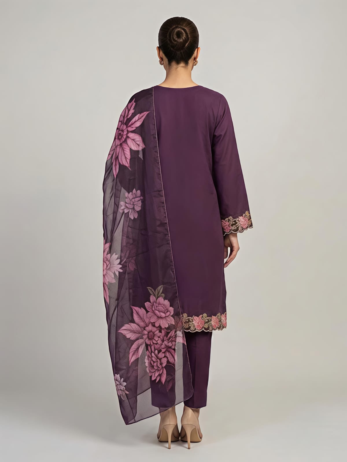 A woman modeling a deep purple three-piece traditional outfit with floral embroidery and a matching sheer dupatta, set against a plain light gray studio background.