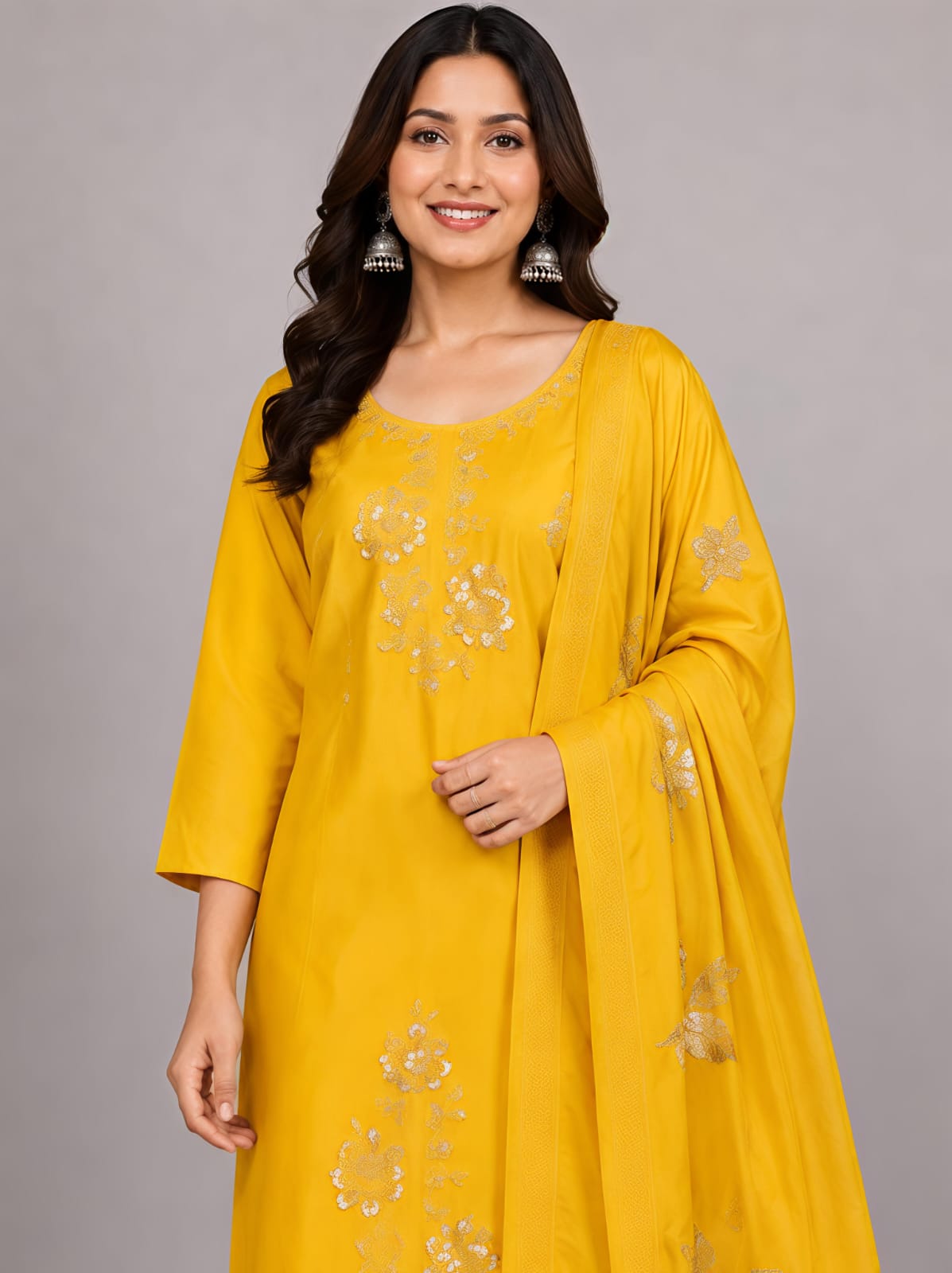 A woman modeling a bright yellow traditional Indian outfit with matching pants and a sheer dupatta, both decorated with gold floral embroidery, against a solid gray studio background.