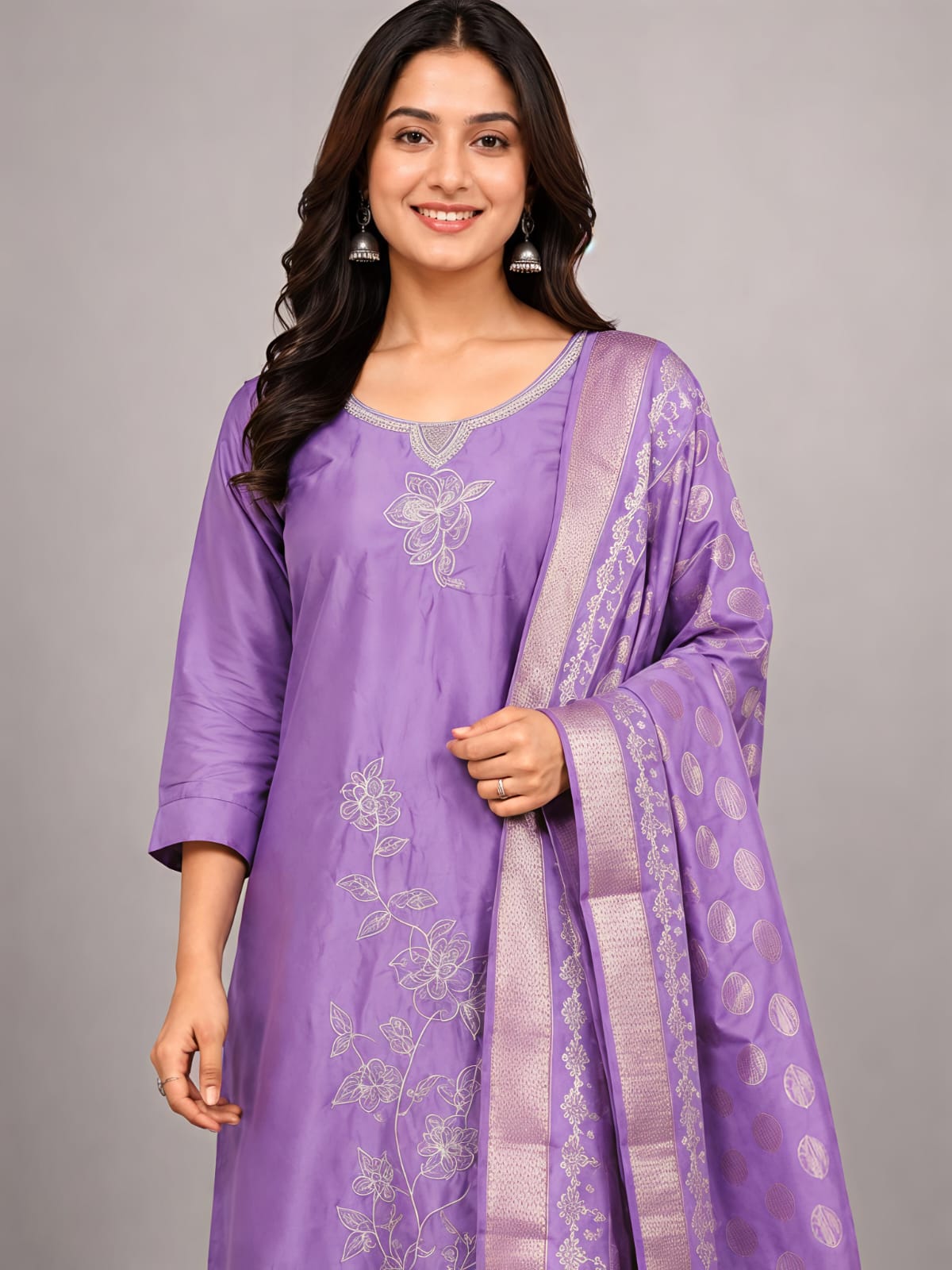 A woman modeling a lavender purple traditional three-piece suit with silver embroidery and a matching dupatta, set against a plain gray studio background.