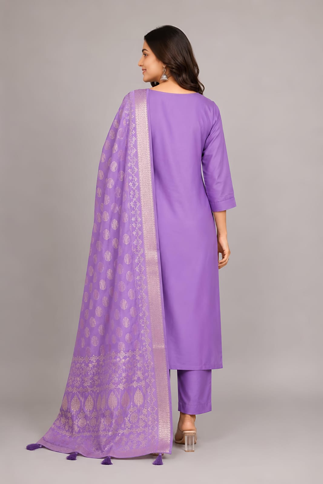 A woman modeling a lavender purple traditional three-piece suit with silver embroidery and a matching dupatta, set against a plain gray studio background.