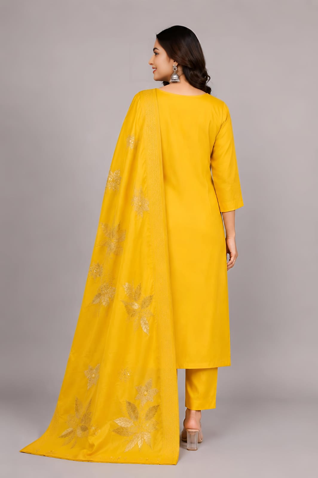 A woman modeling a bright yellow traditional Indian outfit with matching pants and a sheer dupatta, both decorated with gold floral embroidery, against a solid gray studio background.