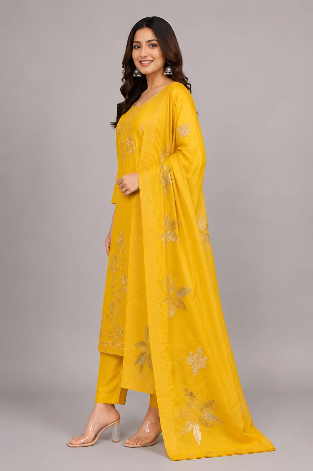 A woman modeling a bright yellow traditional Indian outfit with matching pants and a sheer dupatta, both decorated with gold floral embroidery, against a solid gray studio background.
