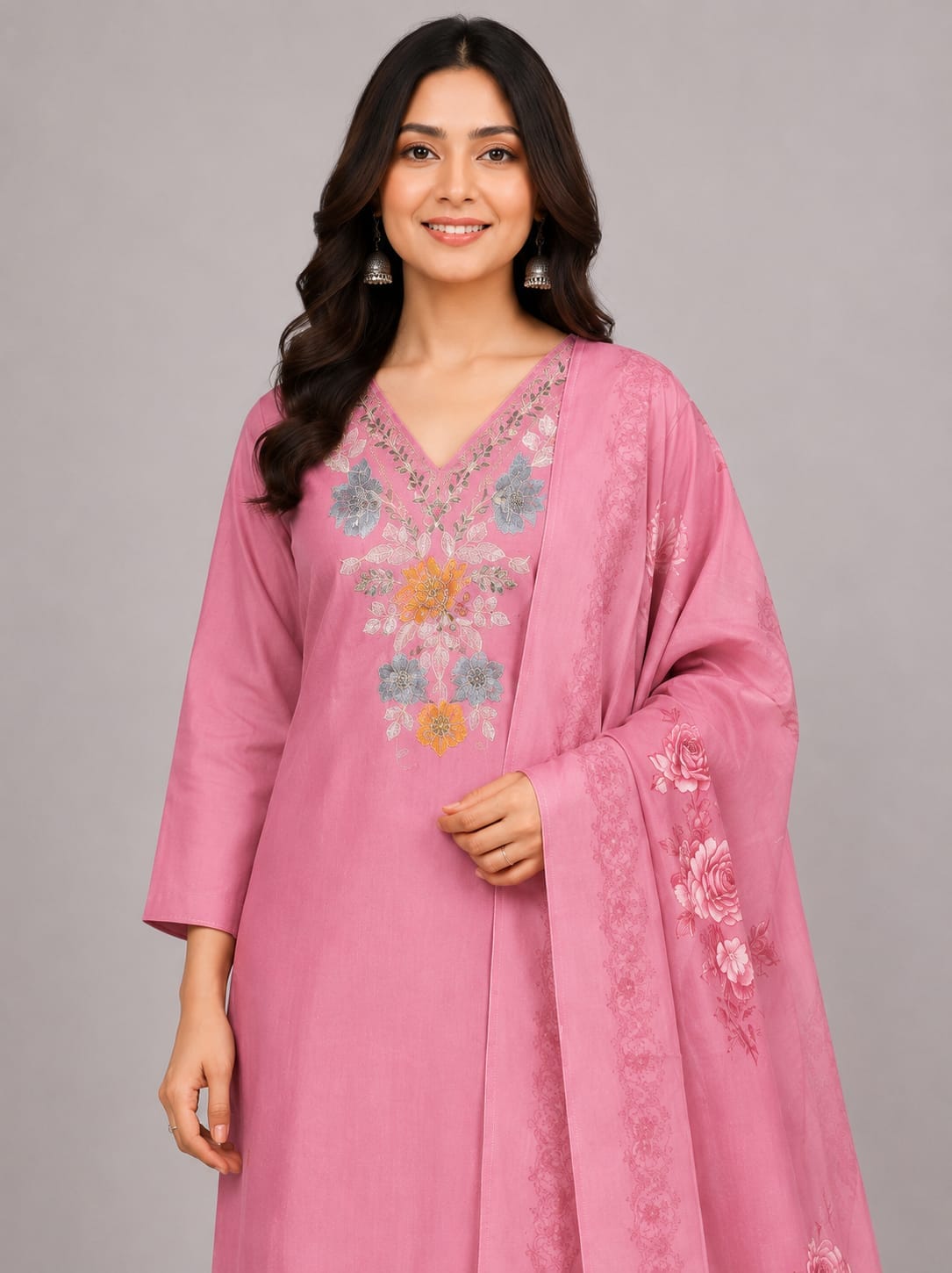 A woman modeling a pink traditional outfit with matching pants and a long sheer dupatta, featuring colorful floral embroidery on the neckline, hem, and dupatta, against a solid gray studio background.