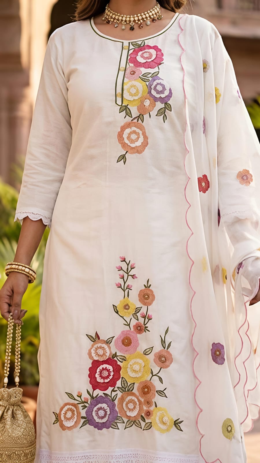 A woman models a white ethnic salwar kameez suit with colorful floral embroidery, matching wide-leg trousers, and a sheer embroidered dupatta, set against an outdoor architectural background with potted plants.