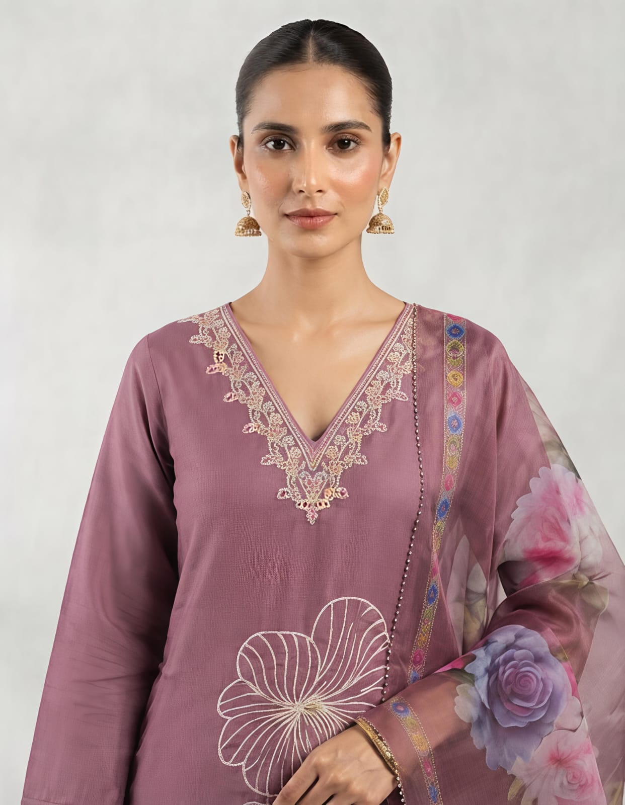 A woman modeling a three-piece traditional outfit consisting of a dusty purple kurta with white floral embroidery, matching trousers, and a sheer floral dupatta, set against a neutral studio backdrop.