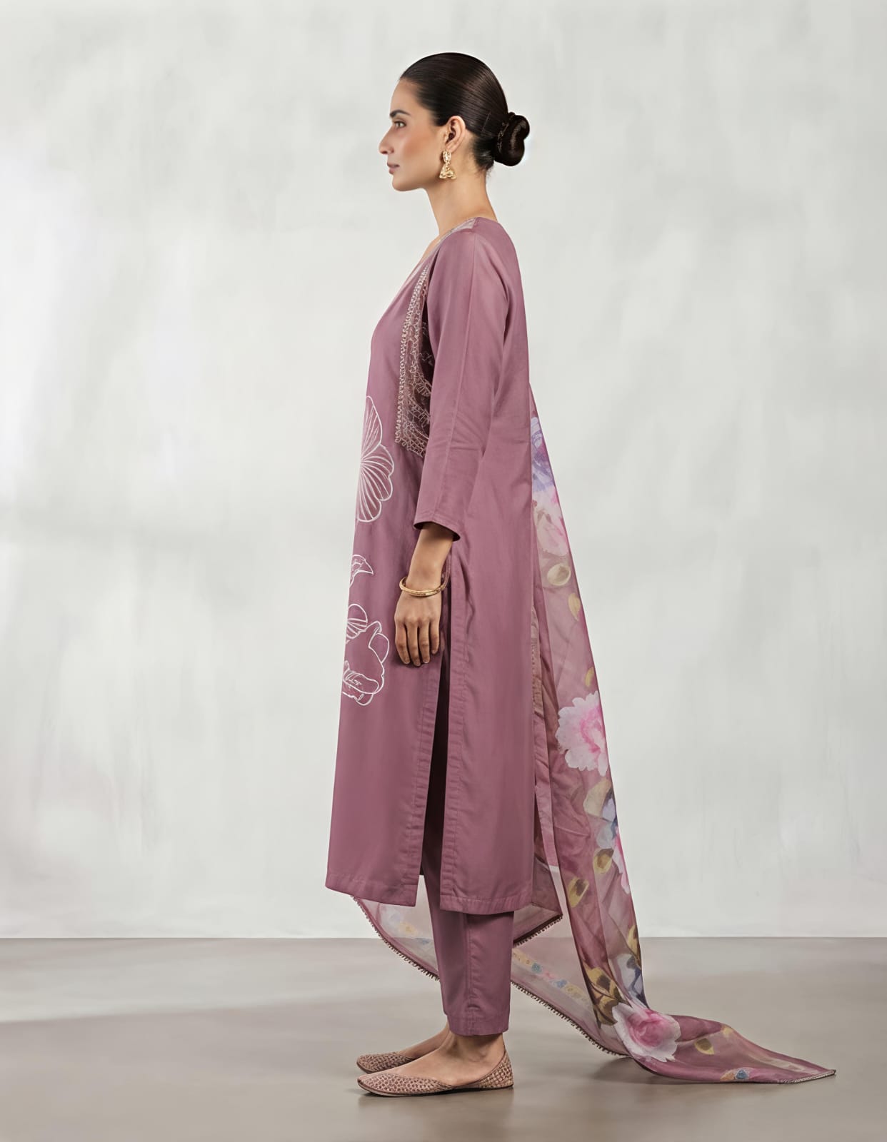 A woman modeling a three-piece traditional outfit consisting of a dusty purple kurta with white floral embroidery, matching trousers, and a sheer floral dupatta, set against a neutral studio backdrop.