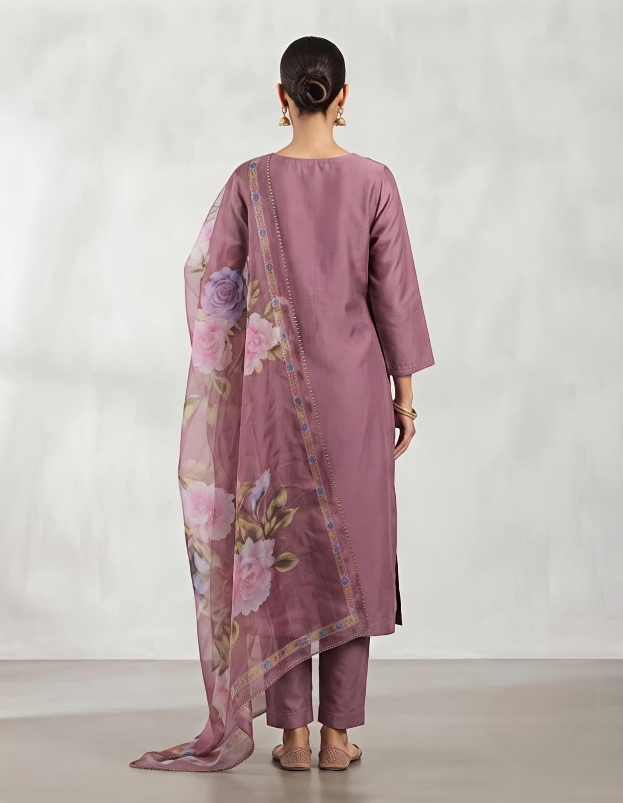 A woman modeling a three-piece traditional outfit consisting of a dusty purple kurta with white floral embroidery, matching trousers, and a sheer floral dupatta, set against a neutral studio backdrop.