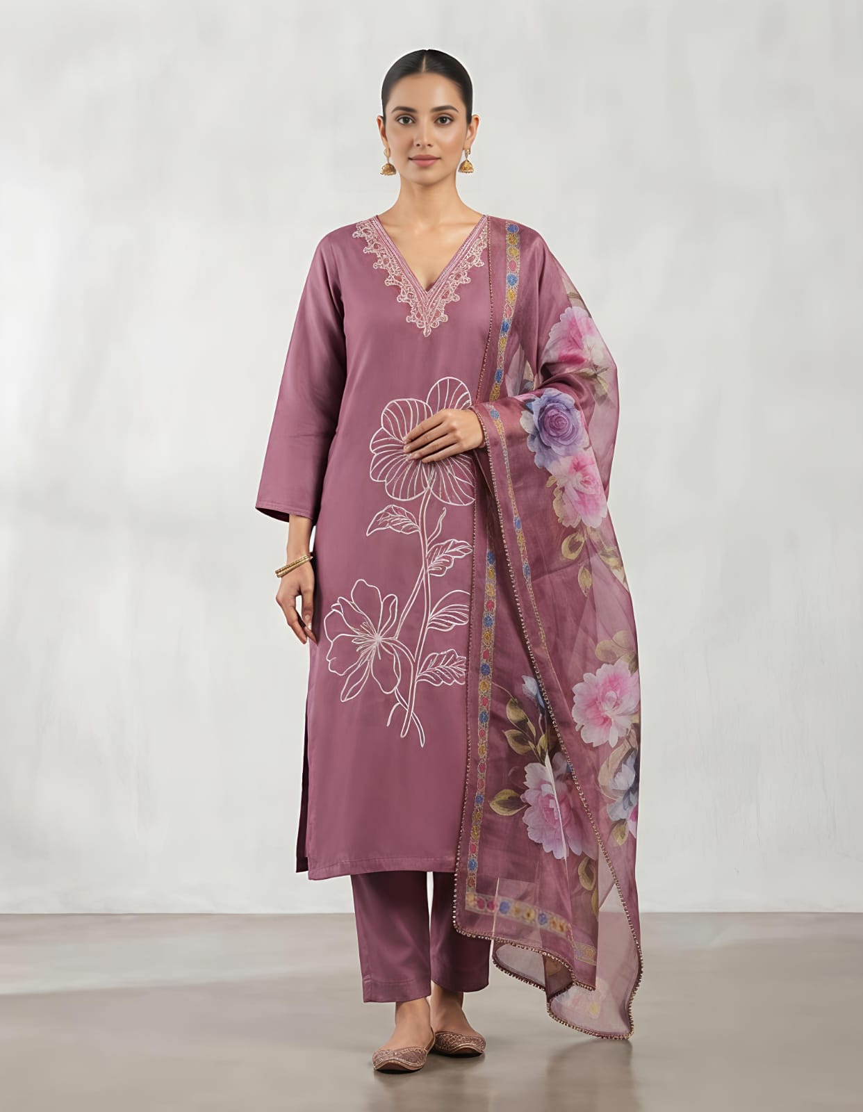 A woman modeling a three-piece traditional outfit consisting of a dusty purple kurta with white floral embroidery, matching trousers, and a sheer floral dupatta, set against a neutral studio backdrop.