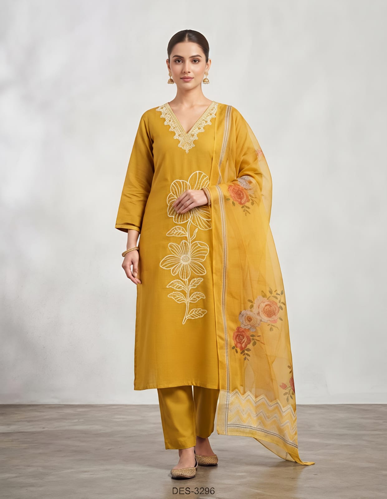 A woman modeling a mustard yellow ethnic suit set featuring a long kurta with white floral embroidery, matching trousers, and a sheer dupatta with orange and yellow floral prints, set against a neutral studio backdrop.