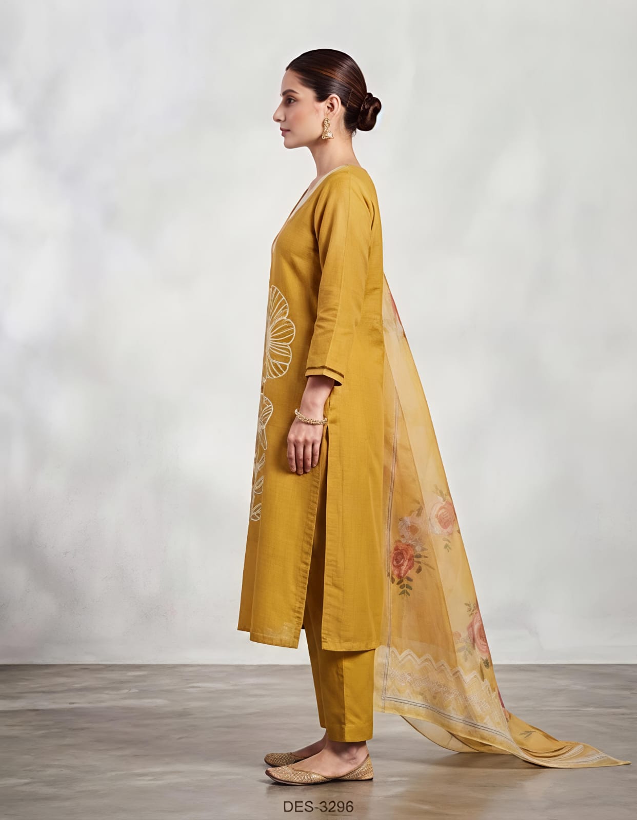 A woman modeling a mustard yellow ethnic suit set featuring a long kurta with white floral embroidery, matching trousers, and a sheer dupatta with orange and yellow floral prints, set against a neutral studio backdrop.