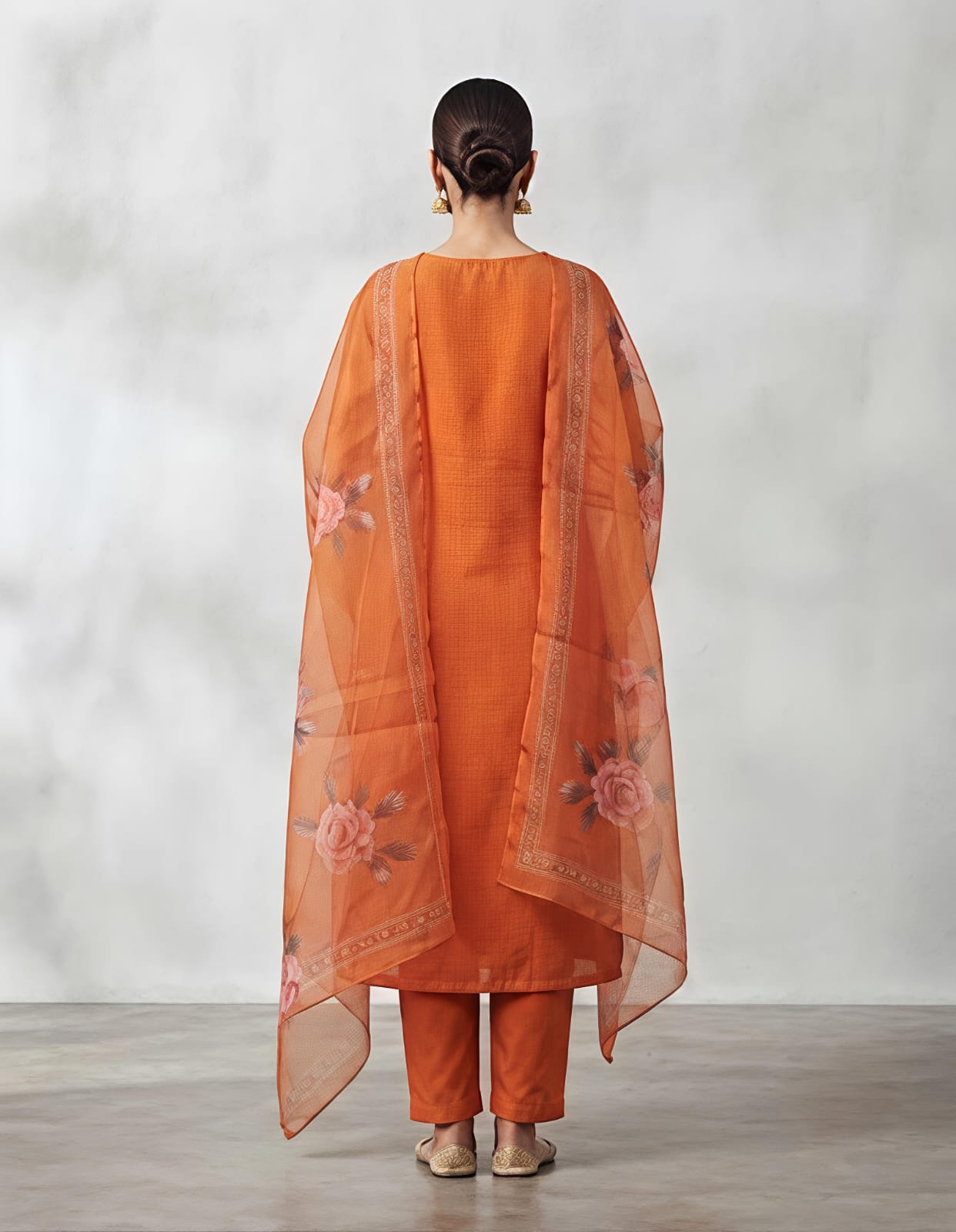 A woman modeling a traditional Indian salwar kameez suit in a vibrant orange color, featuring a kurta with intricate gold and silver embroidery on the chest, matching orange trousers, and a sheer orange dupatta with floral prints and border detailing, set against a neutral gray studio background.