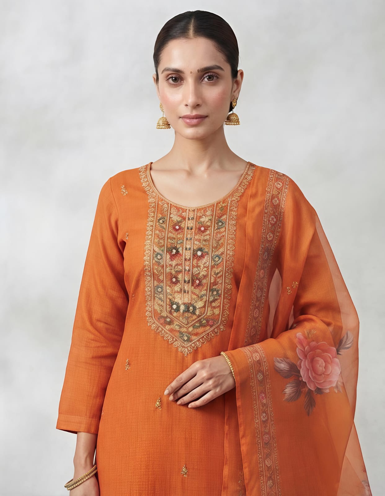 A woman modeling a traditional Indian salwar kameez suit in a vibrant orange color, featuring a kurta with intricate gold and silver embroidery on the chest, matching orange trousers, and a sheer orange dupatta with floral prints and border detailing, set against a neutral gray studio background.