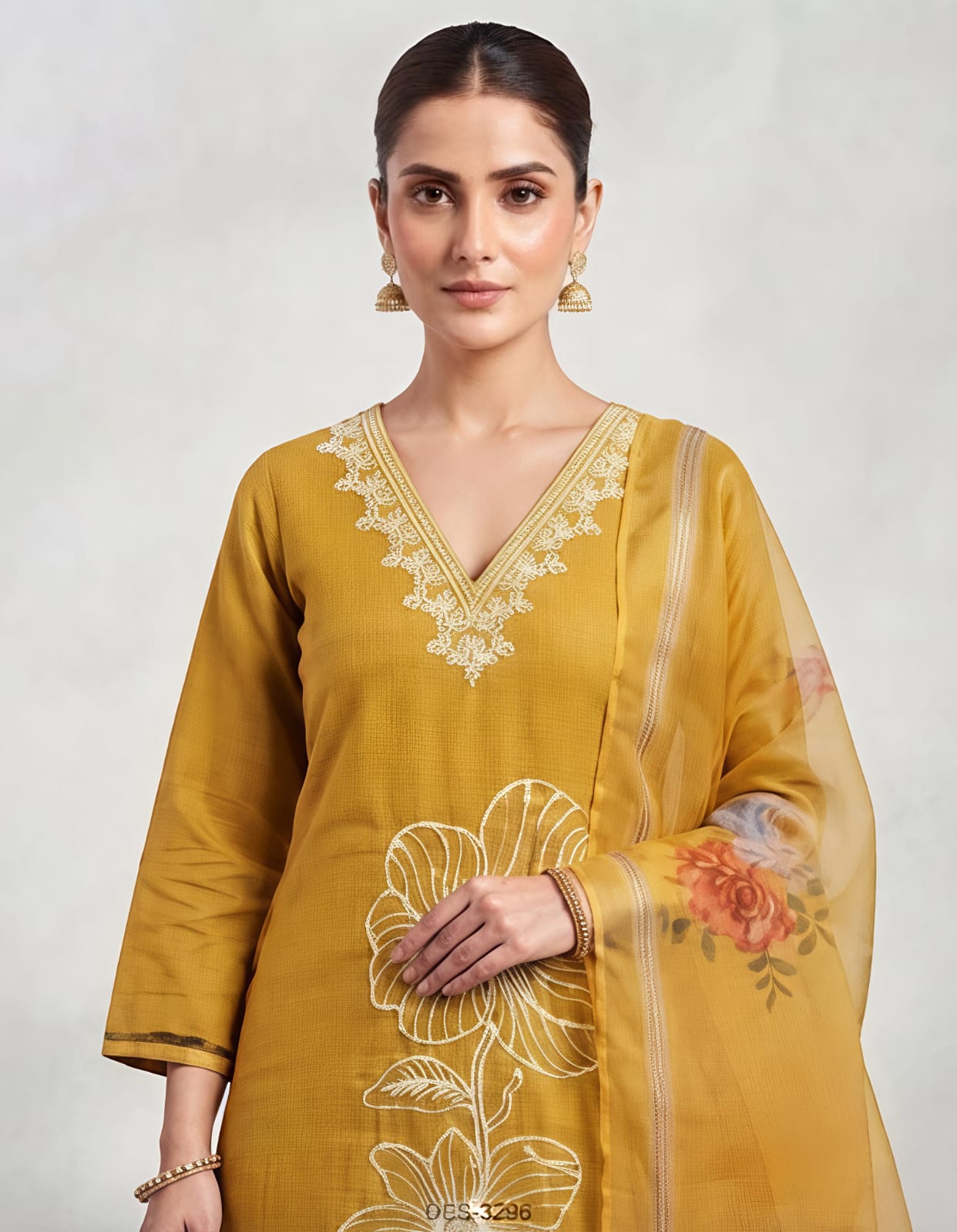 A woman modeling a mustard yellow ethnic suit set featuring a long kurta with white floral embroidery, matching trousers, and a sheer dupatta with orange and yellow floral prints, set against a neutral studio backdrop.