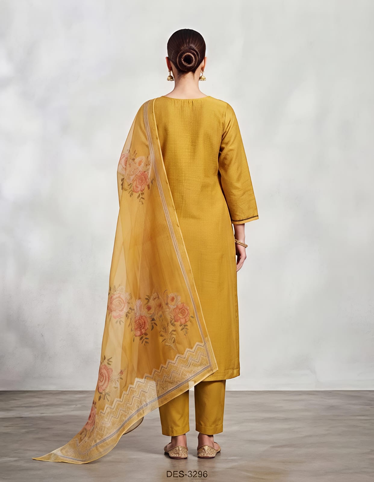 A woman modeling a mustard yellow ethnic suit set featuring a long kurta with white floral embroidery, matching trousers, and a sheer dupatta with orange and yellow floral prints, set against a neutral studio backdrop.