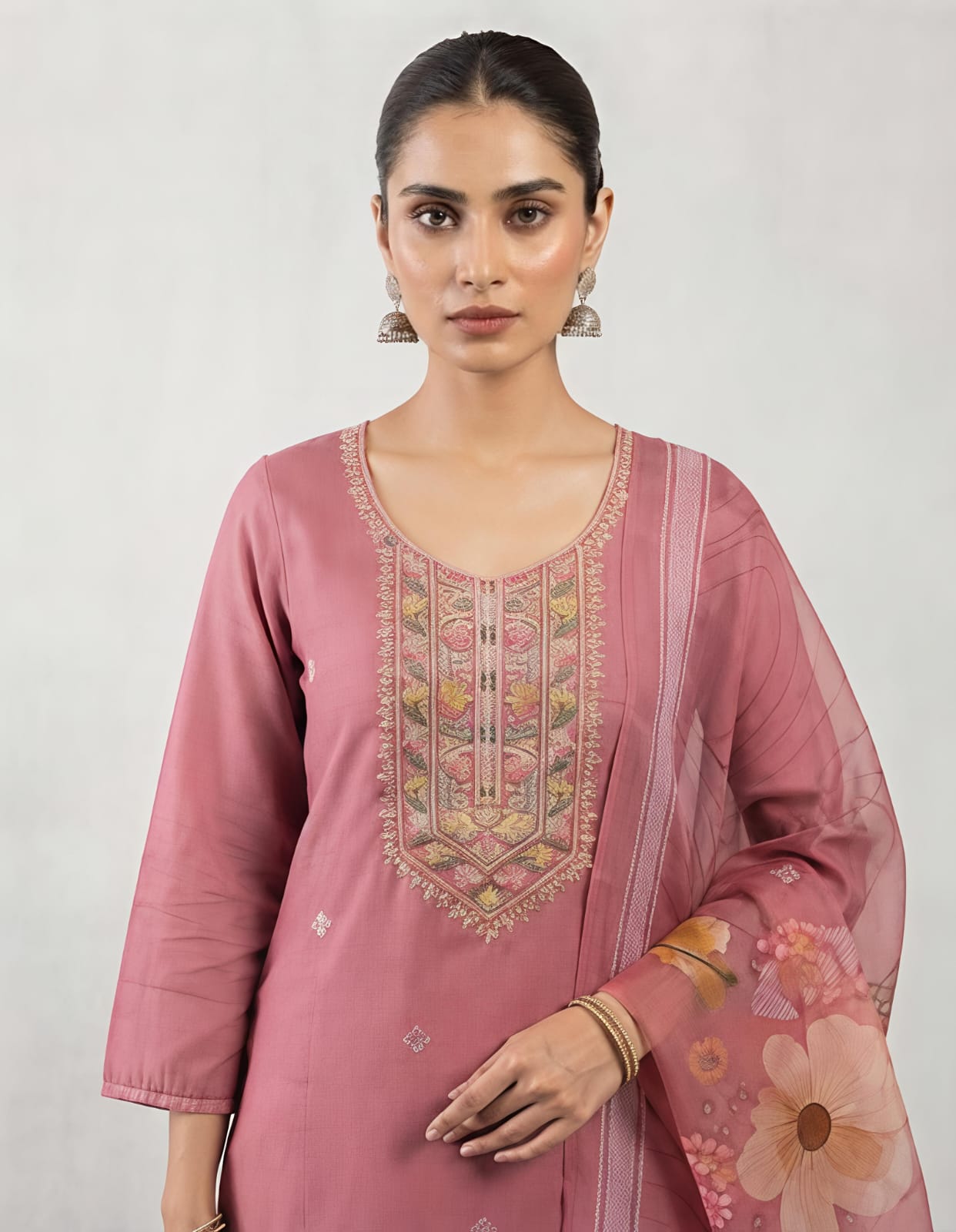 A woman modeling a dusty pink traditional kurta with intricate multicolor embroidery on the chest and a matching sheer floral dupatta, set against a plain light gray background.