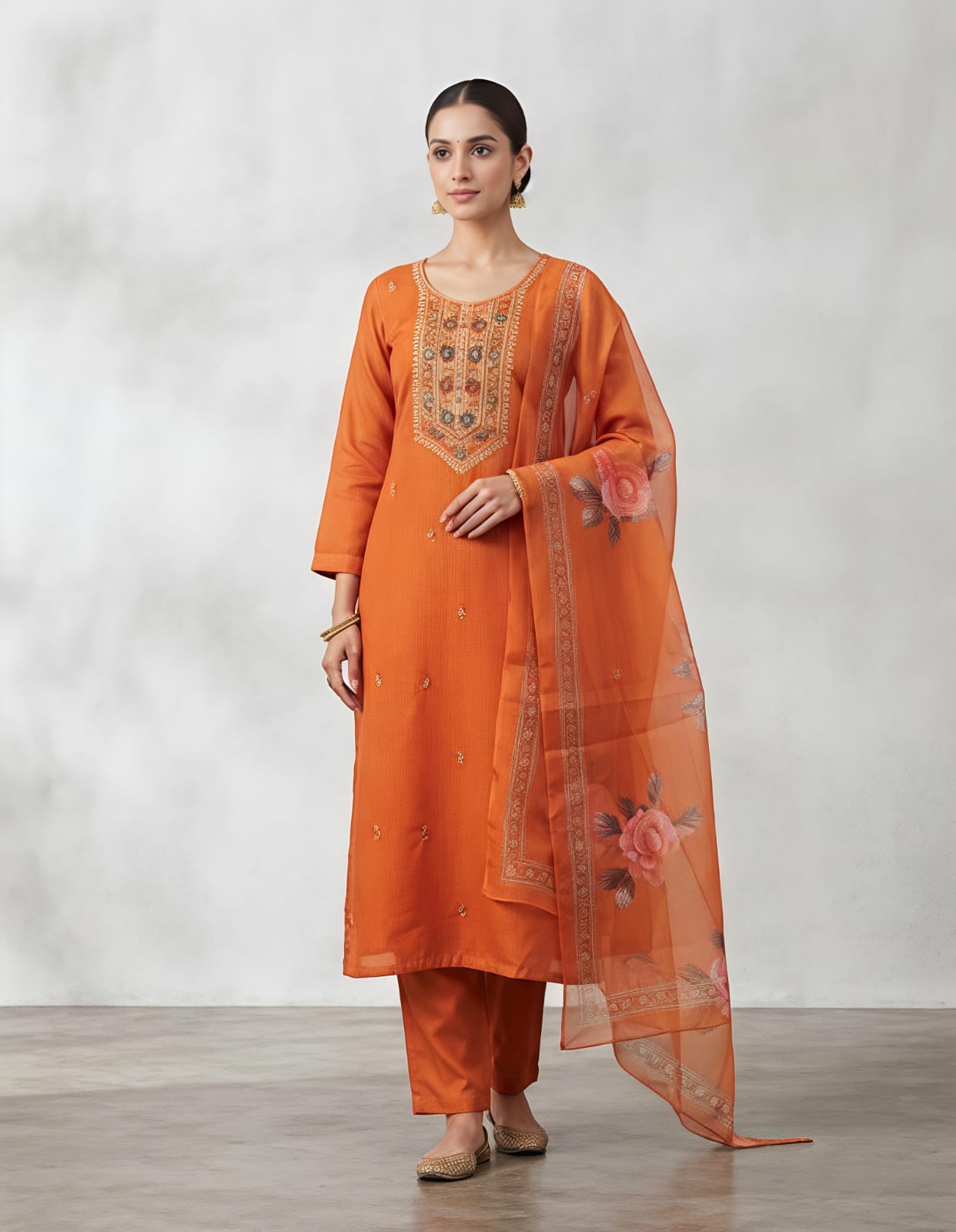 A woman modeling a traditional Indian salwar kameez suit in a vibrant orange color, featuring a kurta with intricate gold and silver embroidery on the chest, matching orange trousers, and a sheer orange dupatta with floral prints and border detailing, set against a neutral gray studio background.