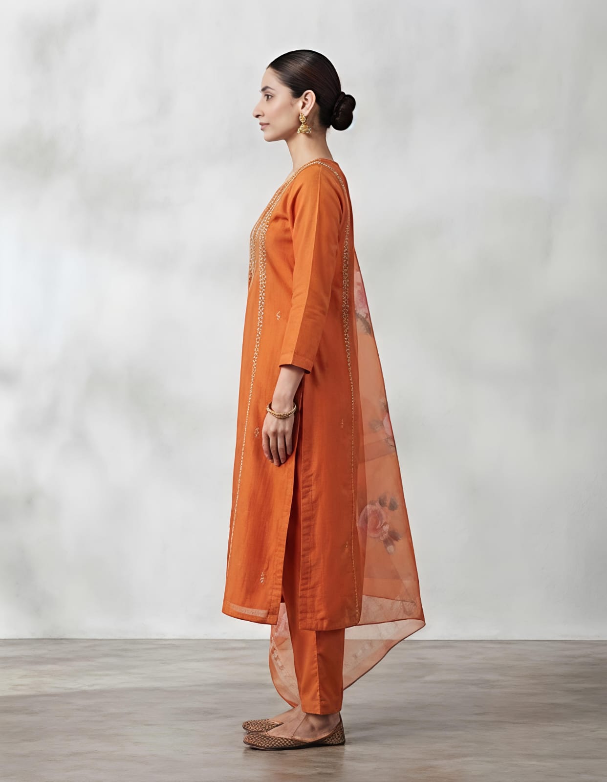 A woman modeling a traditional Indian salwar kameez suit in a vibrant orange color, featuring a kurta with intricate gold and silver embroidery on the chest, matching orange trousers, and a sheer orange dupatta with floral prints and border detailing, set against a neutral gray studio background.