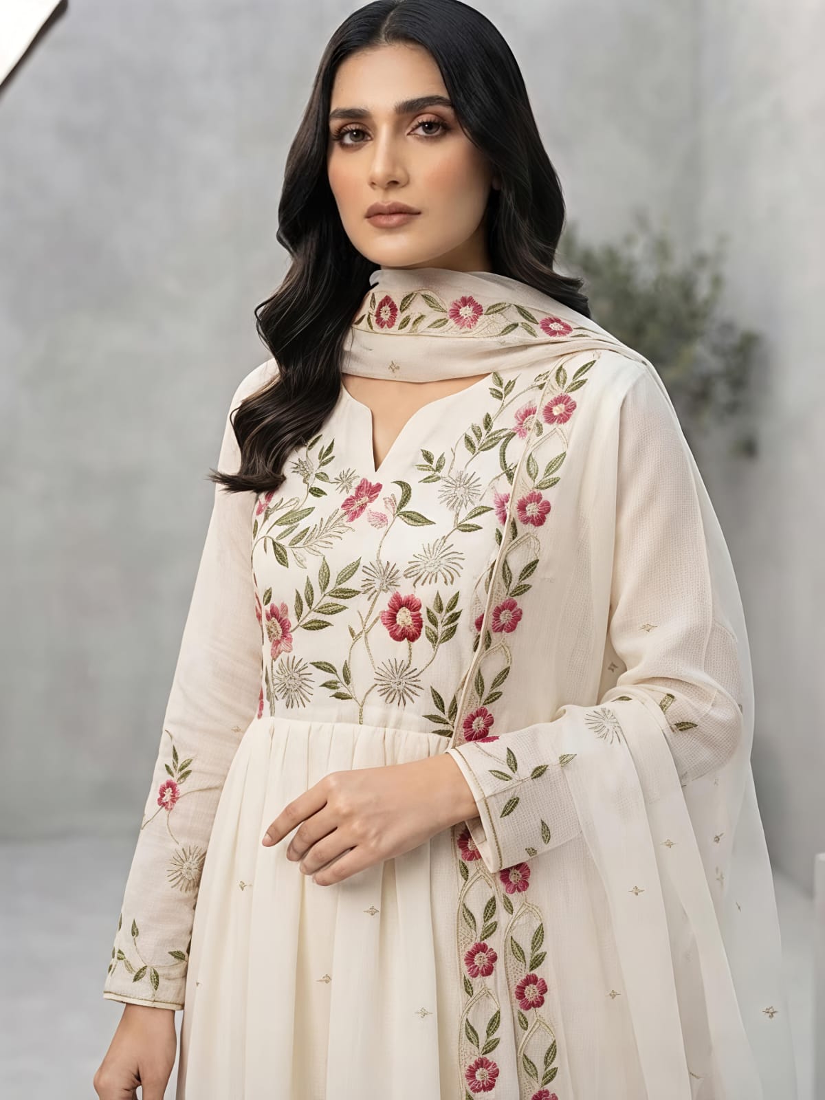 A woman models a traditional Indian outfit consisting of a cream-colored Anarkali kurta with red and green floral embroidery, paired with matching trousers and a sheer dupatta, in a studio setting.